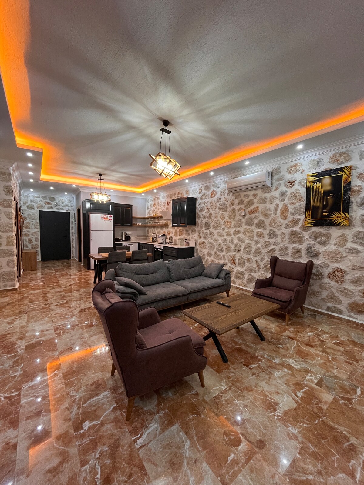 An inviting living area features a gray sofa and two upholstered chairs arranged around a wooden table. Soft lighting complements the natural stone walls, and a kitchen with dark cabinetry can be seen in the background. Glossy tile flooring reflects the ambient light.