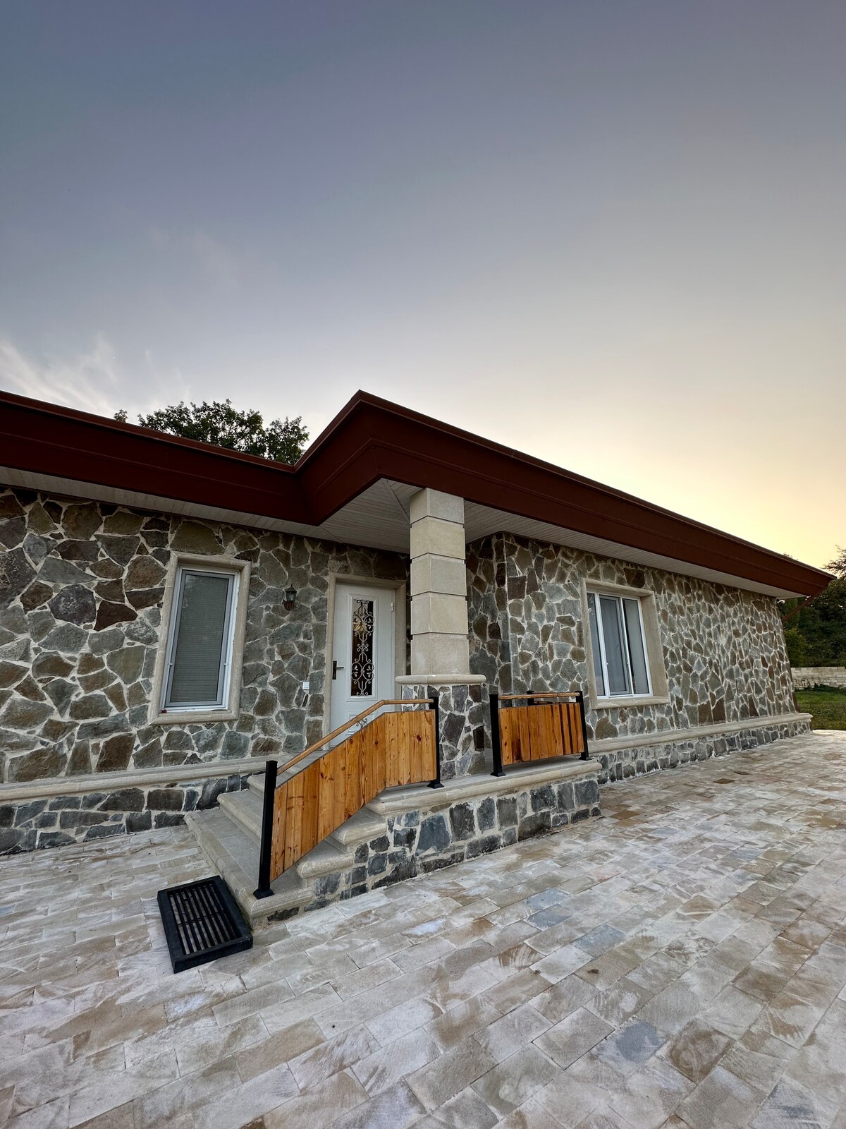 A unique stone house is presented with a combination of light-colored and dark stone exteriors. An accessible wooden ramp leads to the entrance, framed by large windows on either side. The structure is set against a softly lit sky, indicating a peaceful evening.