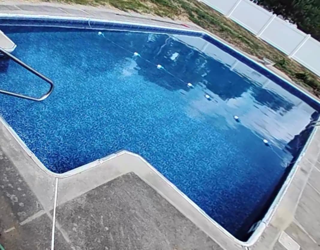 A sparkling, in-ground pool is set against a clean concrete deck. The water shimmers in various shades of blue, while floating markers delineate lanes within the pool. The surrounding area is enclosed by a white fence and features a grassy border.