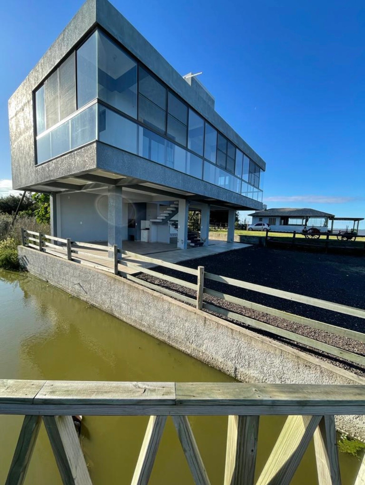 A modern, elevated structure is positioned next to a body of water, featuring large glass windows that allow natural light to illuminate the interior. A wooden railing borders the front area, providing access to the water, while a covered area is visible in the background.