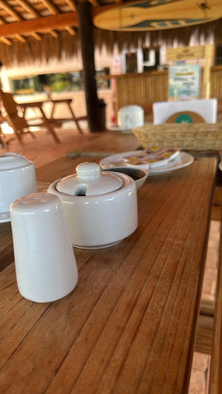 A wooden table is set with white ceramic containers for sugar and salt, alongside a woven basket and a plate with an assortment of items. Soft lighting enhances the rustic ambiance of the space, with a blurred view of the surrounding area in the background.