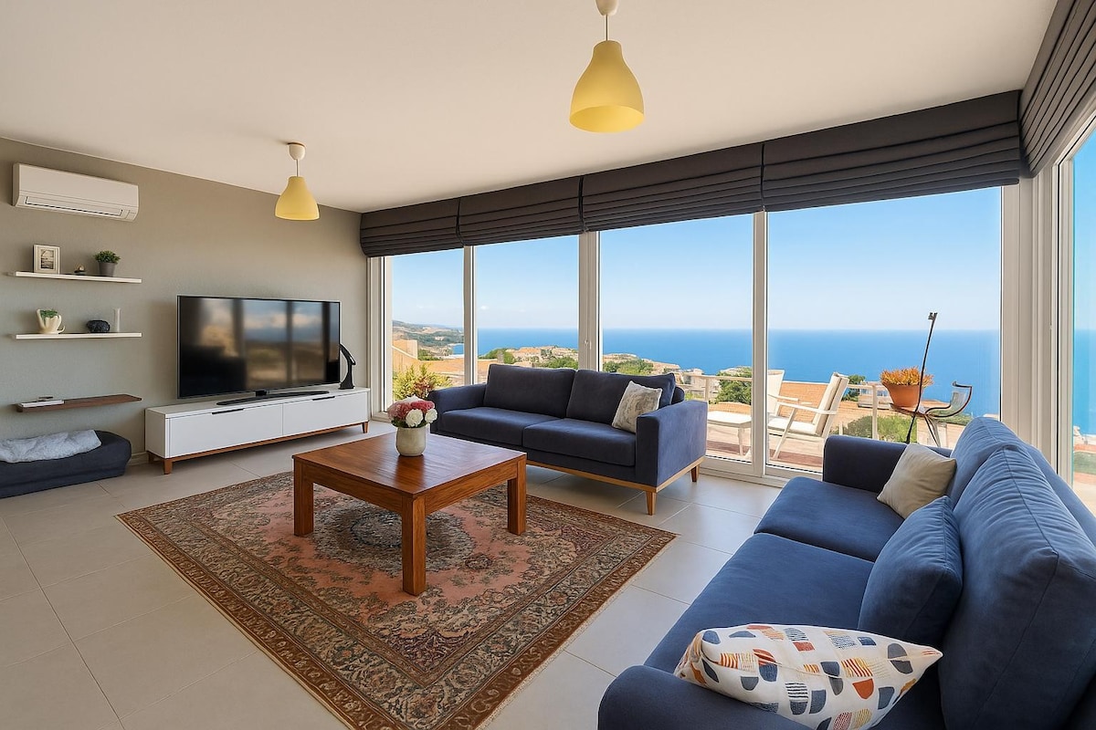 A spacious living room is highlighted by large windows offering ocean views. A wooden coffee table sits at the center, surrounded by two comfortable sofas. A television is mounted on the wall, with a shelf for decorative items and a rug, adding warmth to the space.