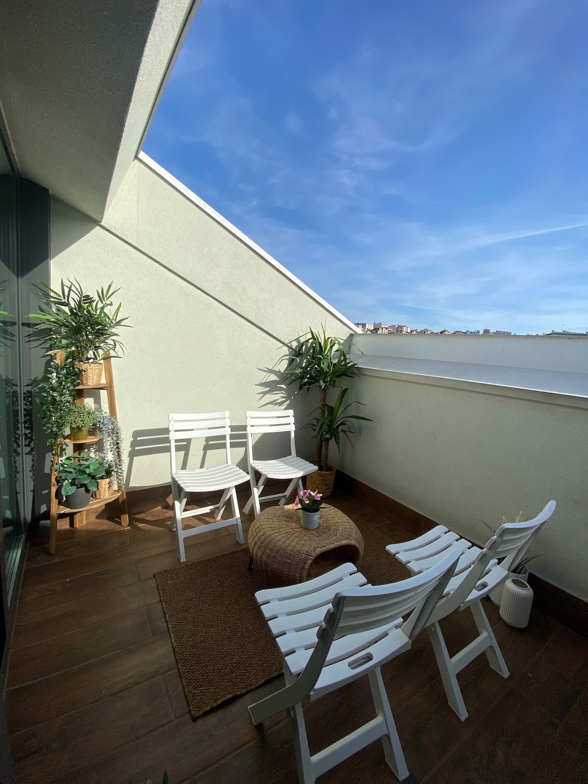 A balcony area is shown, featuring several white chairs arranged around a small round table. Potted plants are positioned in the corner, and natural light illuminates the space through large windows. The flooring is finished in warm wooden tones, creating a comfortable outdoor atmosphere.