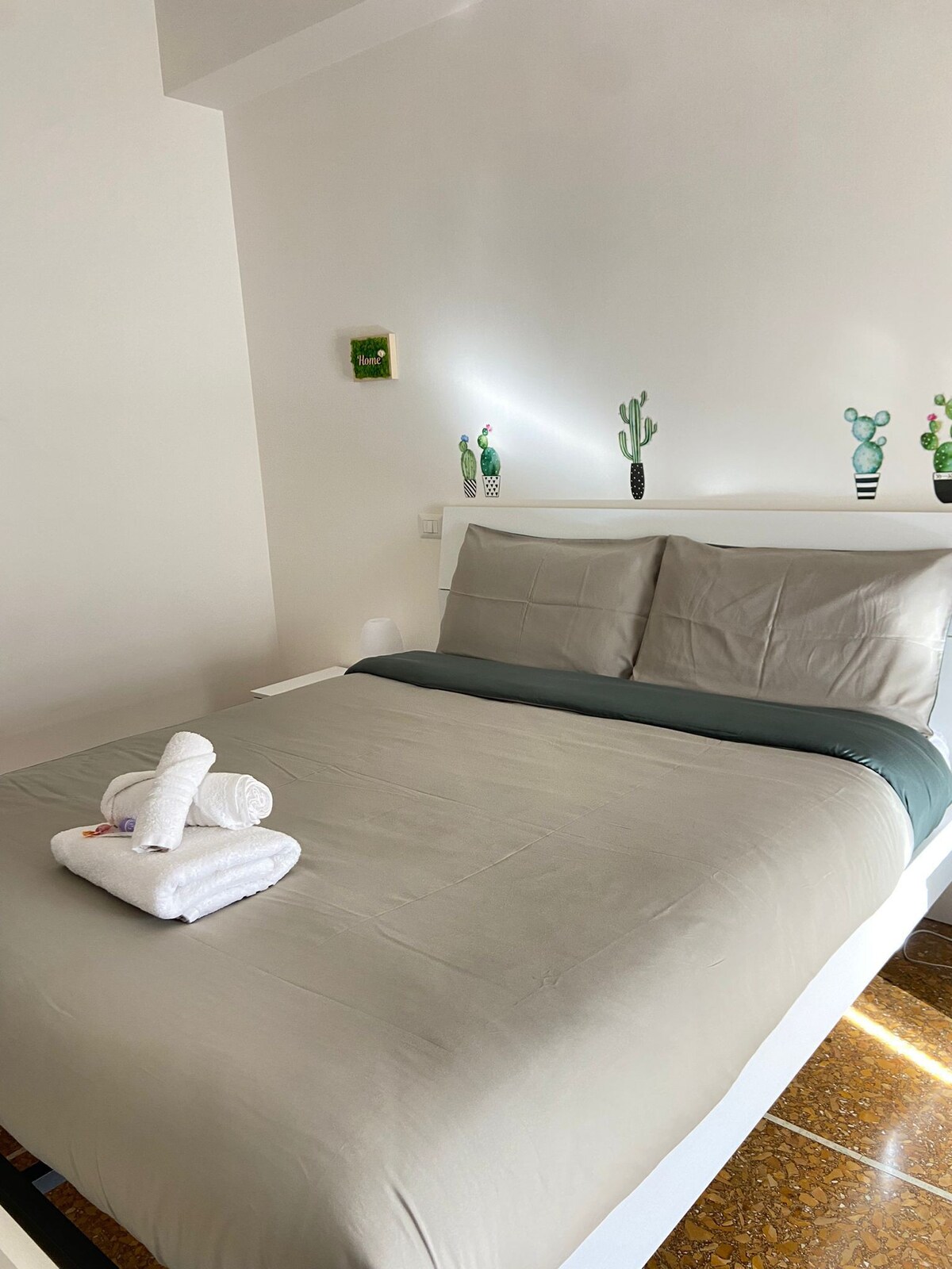 A spacious bed is made with neutral-coloured linens, accented by a contrasting grey throw. White towels are neatly arranged at the foot of the bed. The walls are decorated with artistic cactus designs, contributing to a clean and bright atmosphere.