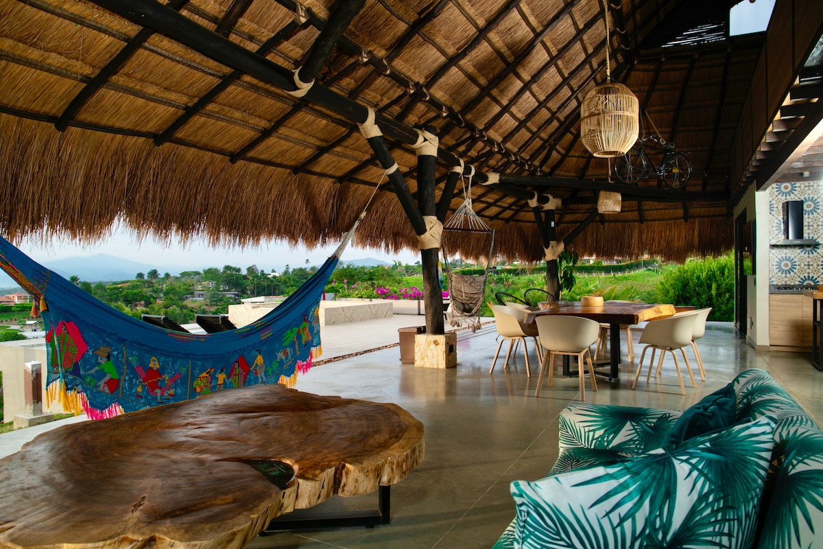 An outdoor seating area is sheltered by a thatched roof, featuring a wooden table surrounded by white chairs. A colorful hammock is displayed, along with a natural wooden coffee table. Lush greenery and distant landscape views are visible in the background.