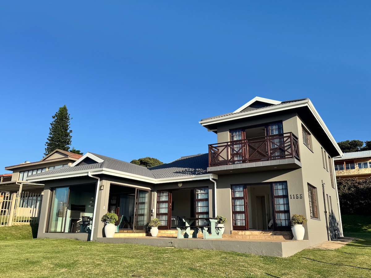 A two-story house is presented with a modern design, featuring large windows and a welcoming porch. Flower pots are placed near the entrance, and a grassy yard extends in front, providing ample outdoor space. The clear blue sky serves as a backdrop.