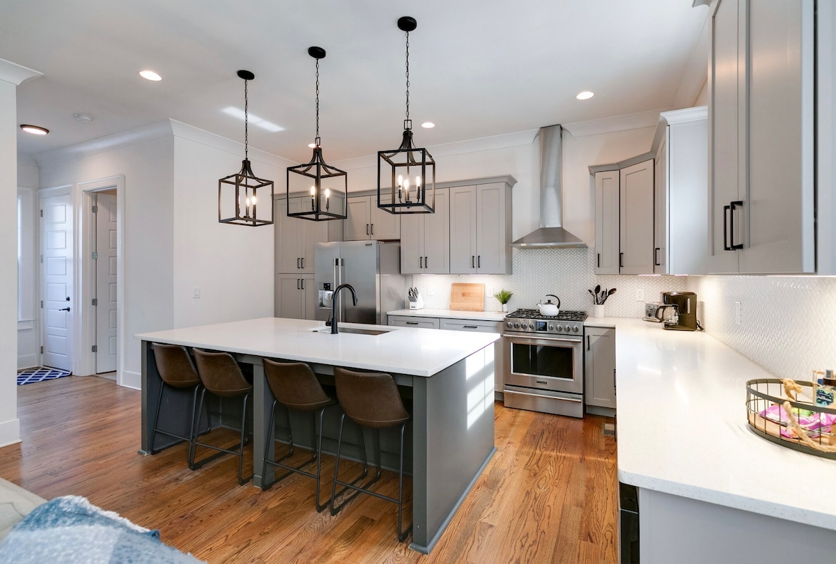 A modern kitchen is highlighted by a large island with four bar stools. Contemporary cabinetry provides ample storage, while stainless steel appliances integrate functionality. Pendant lights hang above the island, illuminating the open space and creating a bright atmosphere.