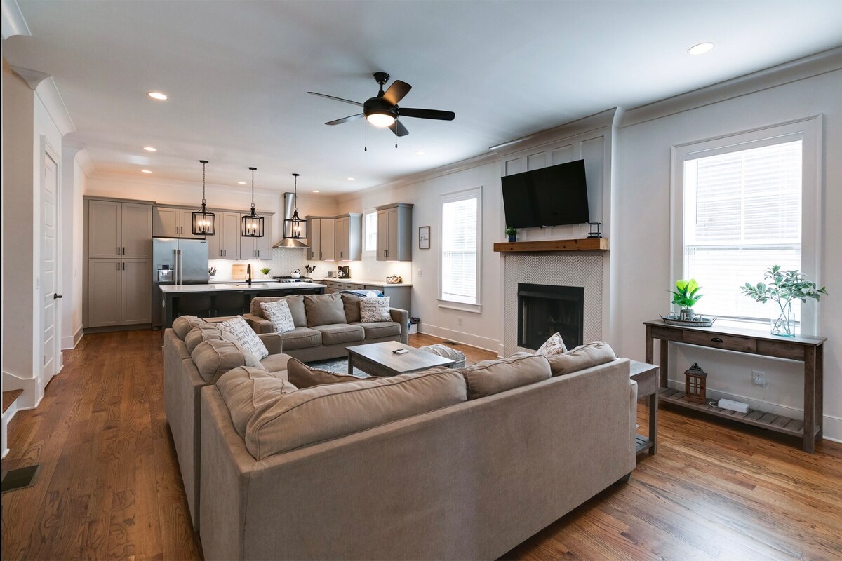 A spacious living area features a light-colored sectional sofa arranged around a coffee table. A mounted television is positioned above a modern fireplace, while the open kitchen with dark cabinetry and pendant lighting is visible in the background. Natural light fills the room through multiple windows.