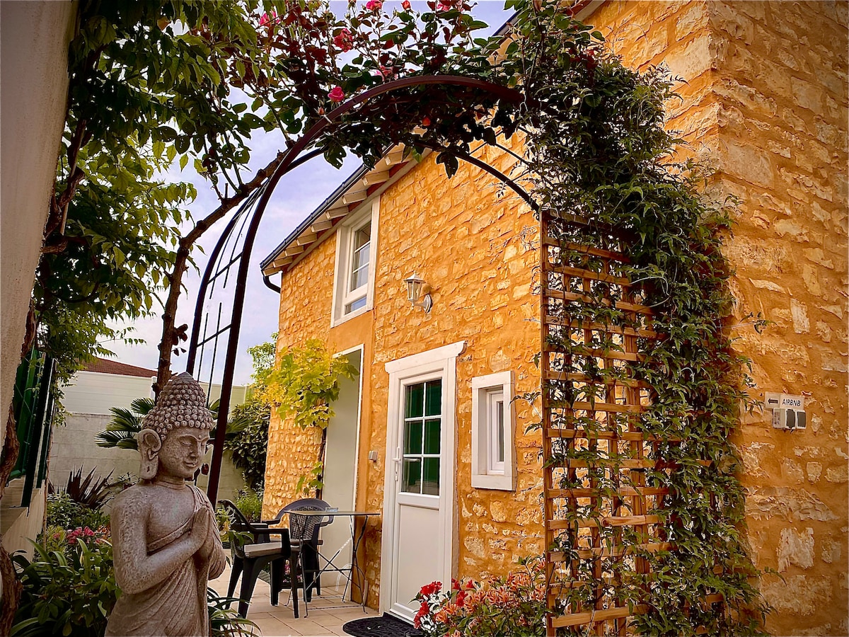 The exterior features a charming stone facade with a vibrant yellow hue. An archway covered in climbing plants leads to a small terrace adorned with a stone Buddha statue. Nearby, a seating area with chairs is visible, surrounded by colorful flowers.