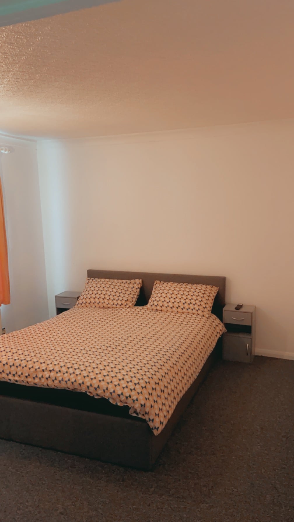 A private bedroom features a spacious bed with a patterned coverlet. Two nightstands flank the bed, and natural light enters through a nearby window covered by a sheer curtain. The room is decorated in neutral tones, providing a calm and simple environment.