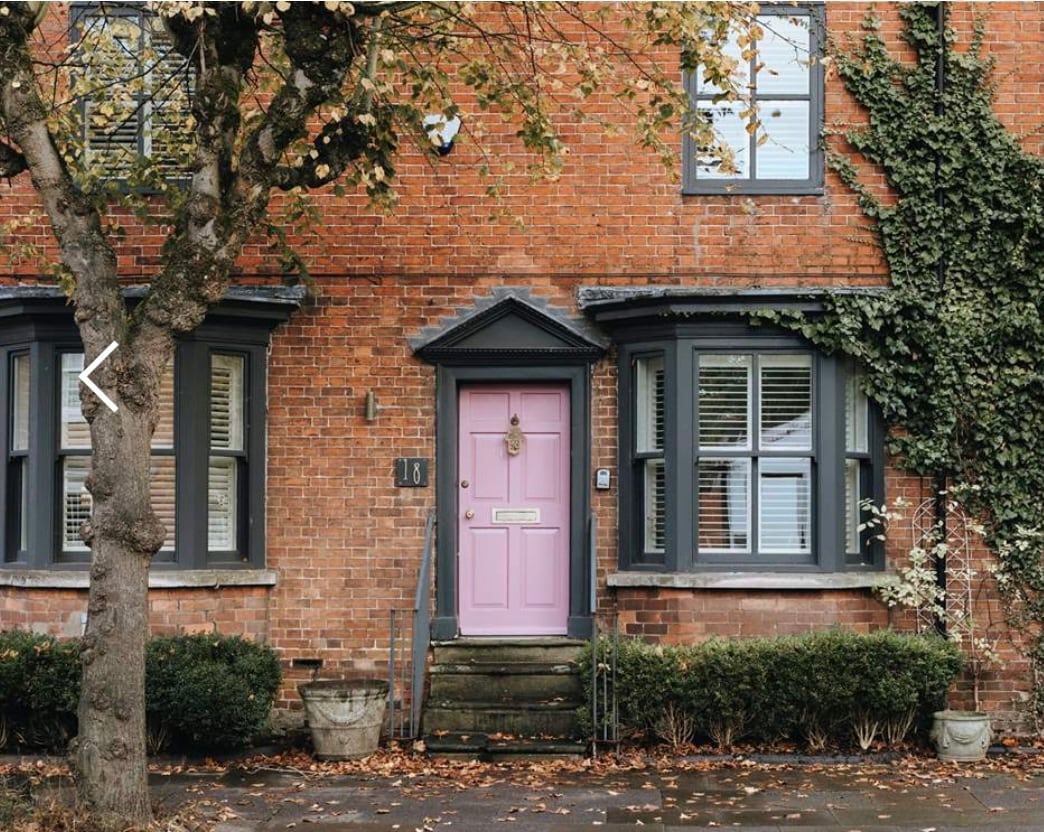 A charming exterior features a pink front door framed by two large windows with shutters. Climbing vines adorn the brick façade, complemented by neatly trimmed hedges and planters at the base of the entry steps. A tree branches gracefully nearby, enhancing the inviting appearance.