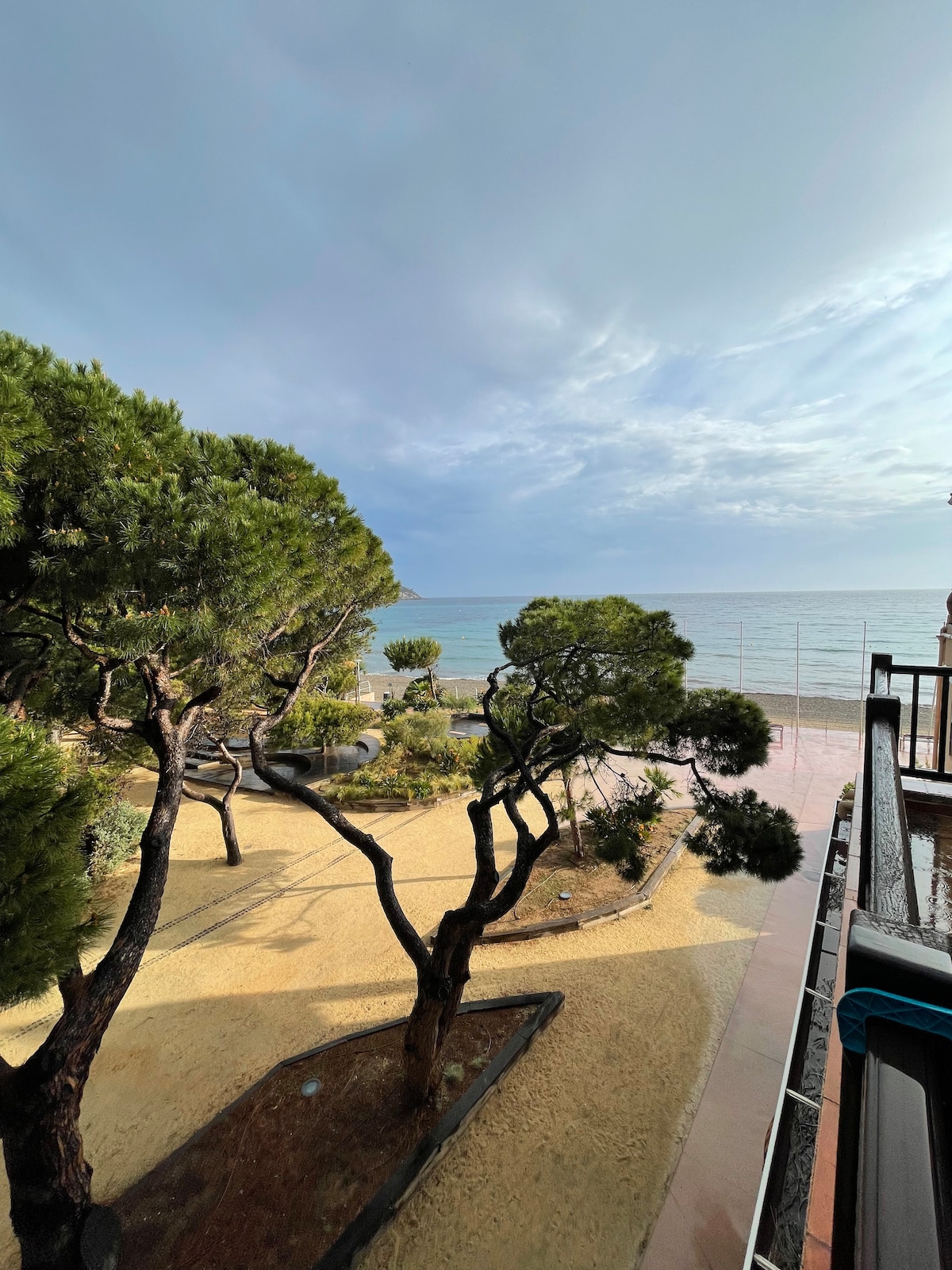 A view of the sandy beach and vibrant sea is framed by trees and greenery. Soft sand paths lead to the water, creating a serene atmosphere. The sky is partially cloudy, allowing natural light to illuminate the landscape.