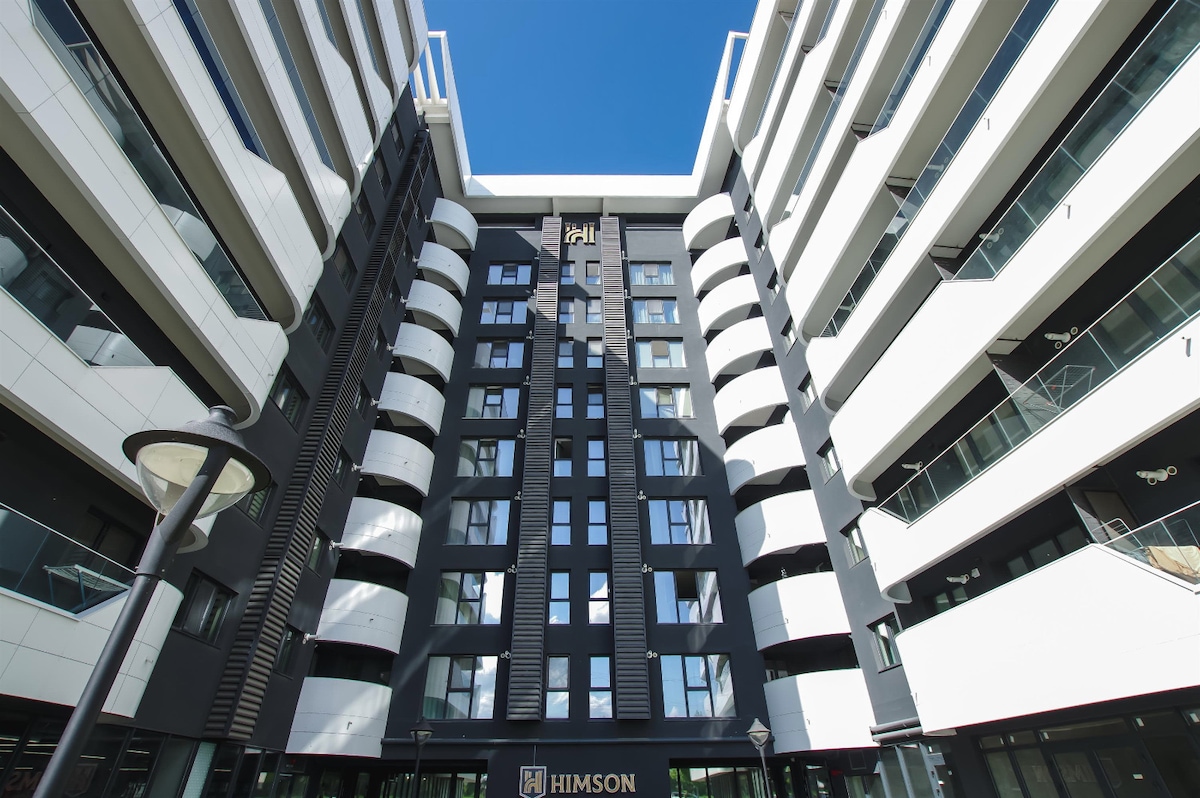 The modern building showcases a unique architectural design, featuring alternating white and dark panels. Large windows are positioned throughout the structure, allowing ample natural light. A prominent sign is displayed at the base, and the clear blue sky serves as a backdrop.