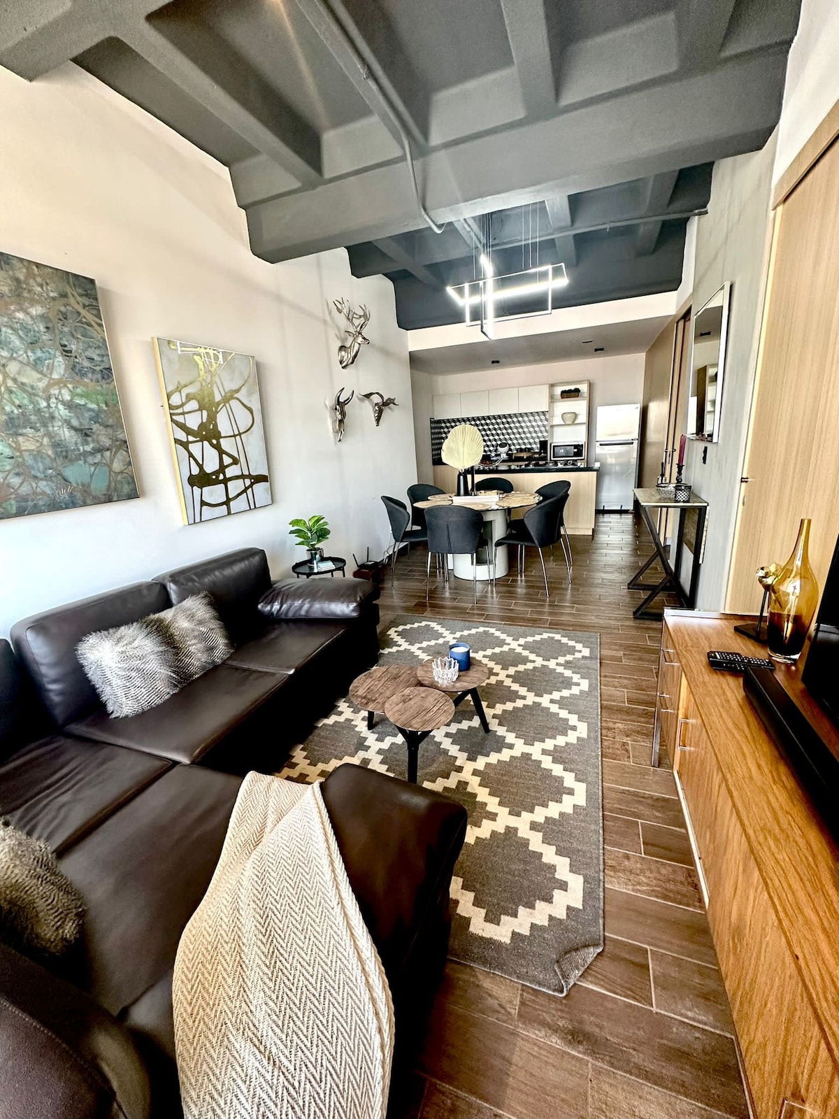 A modern living space features a sectional leather sofa paired with a textured throw and decorative pillows. A geometric rug anchors the seating area, while a dining table with chairs is visible in the background next to a sleek kitchen cabinetry.