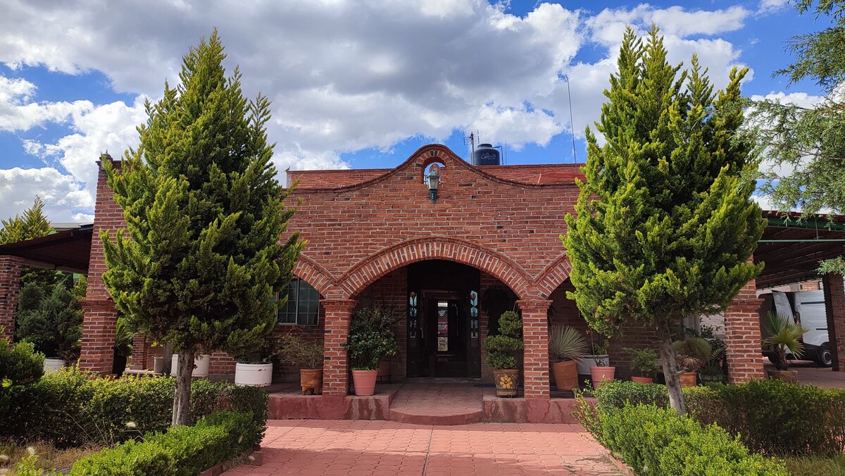 The exterior of a rustic brick building is framed by two tall, leafy conifer trees. Potted plants are arranged along the path leading to the entrance, which features an arched doorway. A partly cloudy sky adds depth to the welcoming facade.