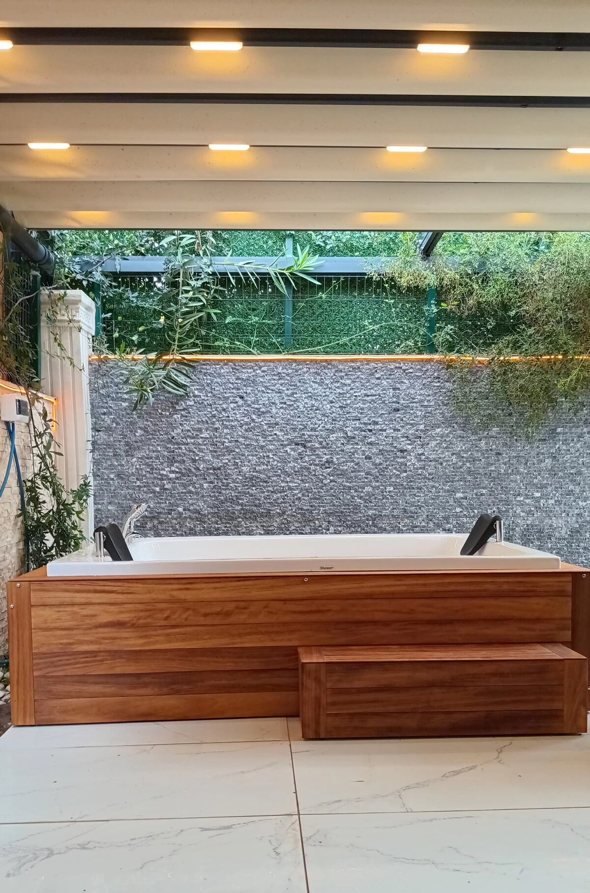 A wooden hot tub is positioned outdoors, featuring a sleek design with two spouts for water. The area is surrounded by a textured stone wall and a greenery backdrop, providing a natural setting. Soft lighting is set above, enhancing the tranquil ambience.
