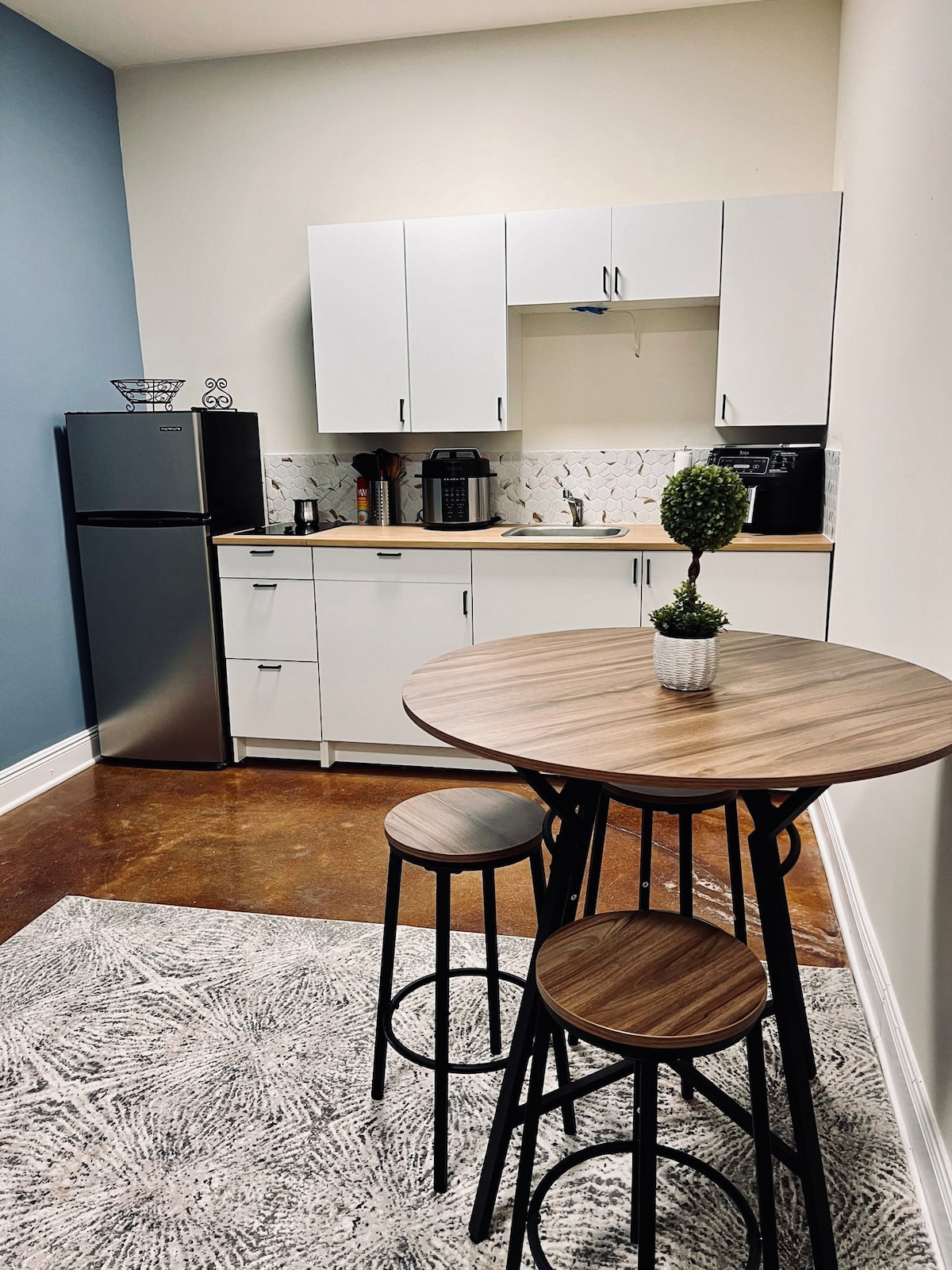 A compact kitchenette is visible, featuring a refrigerator, multiple appliances, and a sink. The space includes a round dining table with three wooden chairs. A small decorative plant sits at the center of the table, and the walls are painted in a light blue tone.