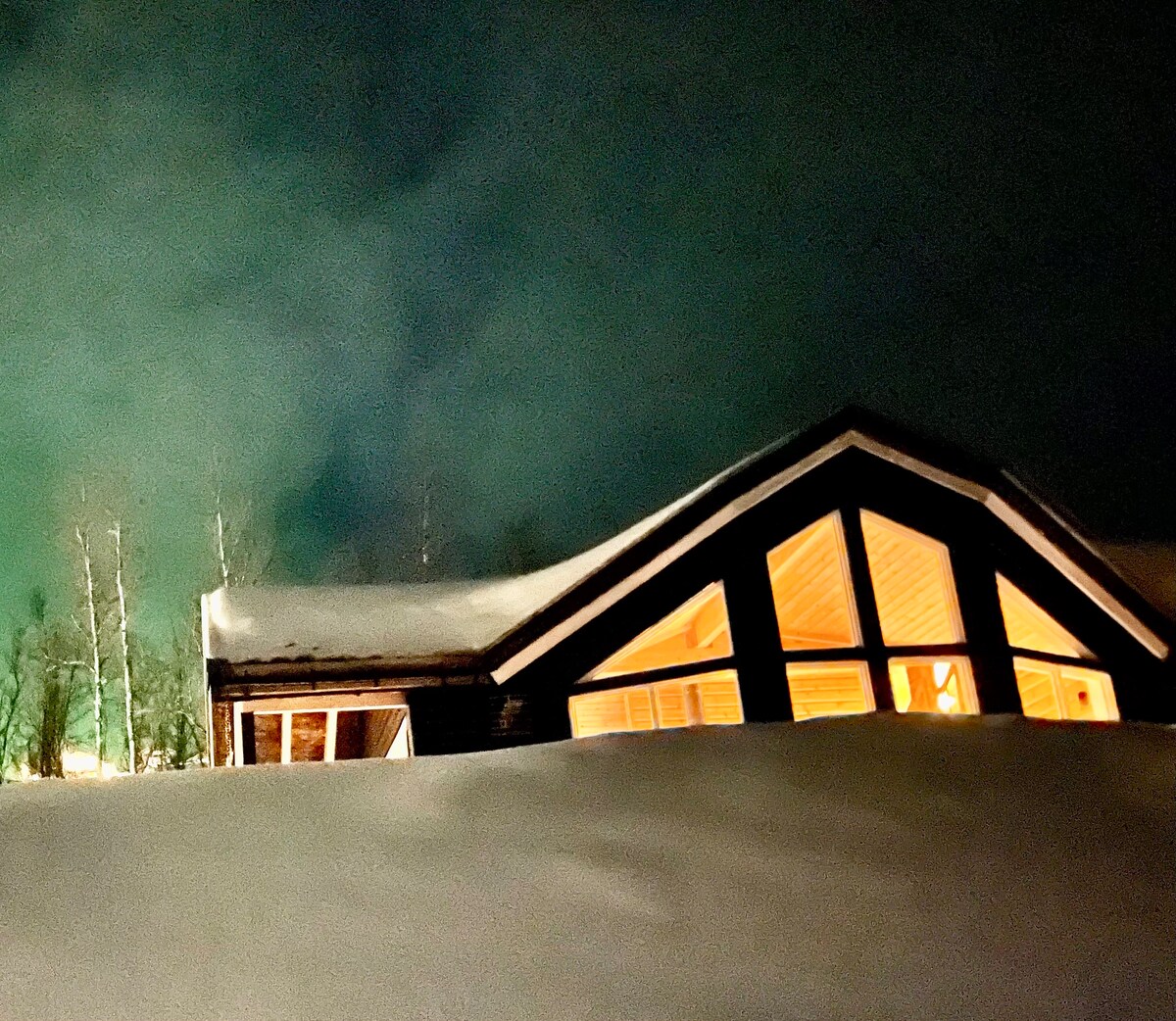 A modern cabin is partially surrounded by snow, with warm light glowing from large windows. The roof is steeply pitched, creating a distinctive silhouette against a night sky that features soft shades of green and blue, hinting at a northern light display.