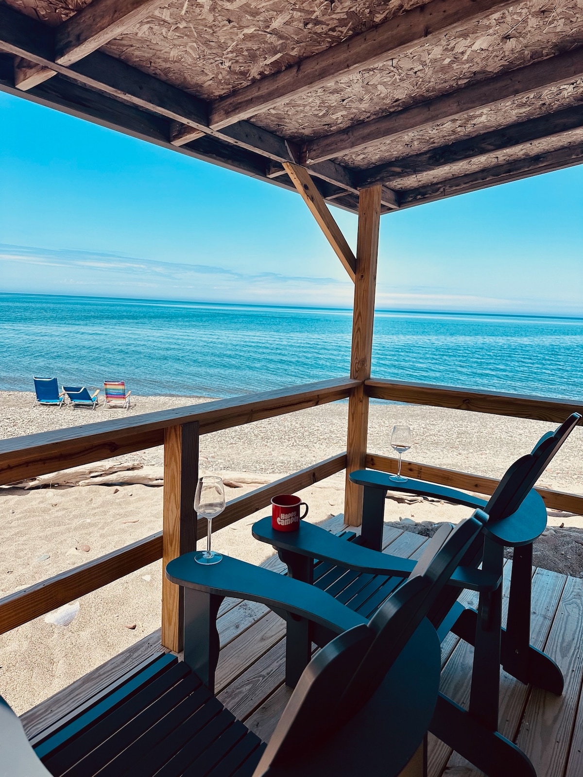 A spacious deck with two dark wooden chairs overlooks a serene beach scene. A cup rests on a small table, and beyond, two colorful lounge chairs are positioned on the sandy shore. The calm waters of Lake Ontario reflect a clear blue sky.