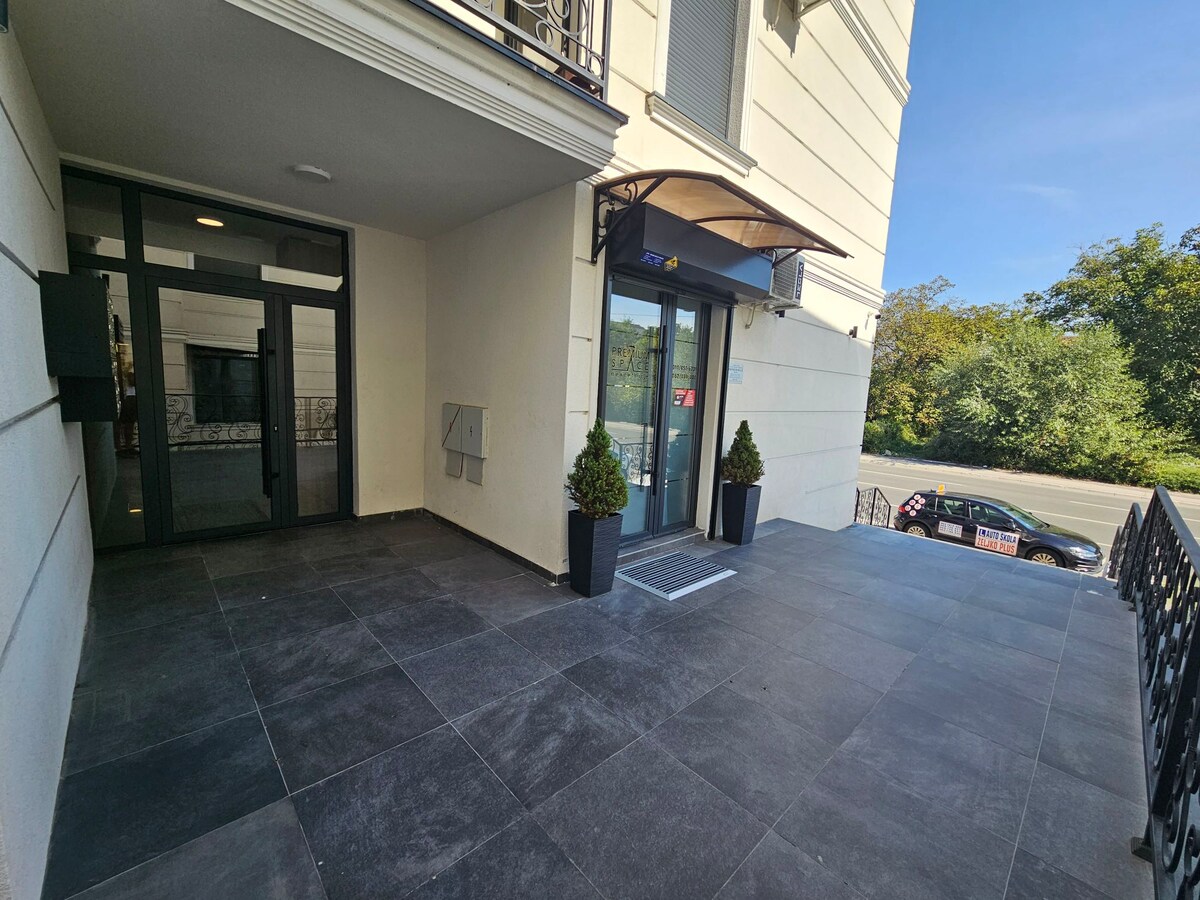 The entrance features a spacious, tiled area framed by large glass doors. Potted plants are positioned near the entrance, and the surrounding landscape includes trees visible in the background. Bright natural light illuminates the welcoming facade.