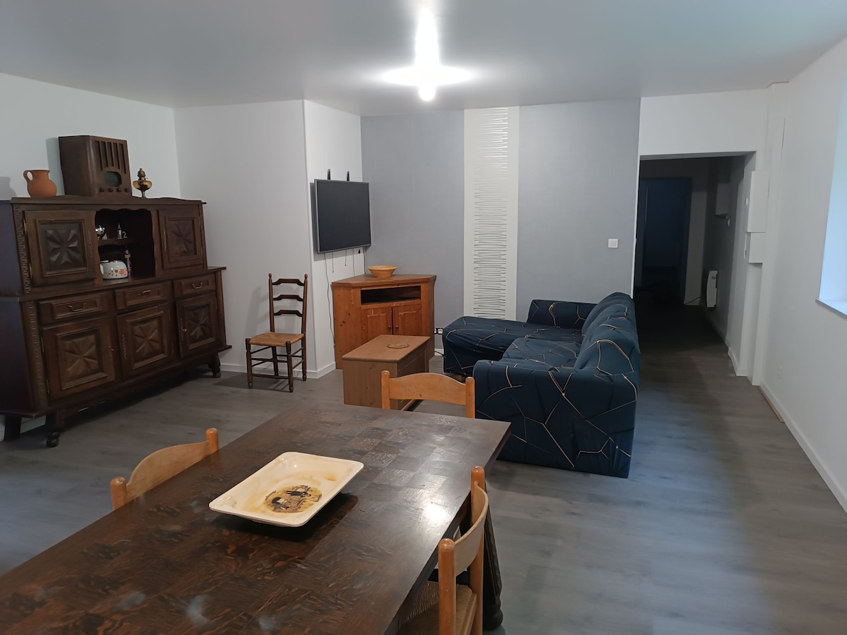 A living area features a comfortable blue sofa with geometric patterns and a wooden coffee table at its center. A dining table with wooden chairs is visible, complemented by a rustic wooden cabinet and a mounted television. Soft lighting brightens the neutral-toned space.