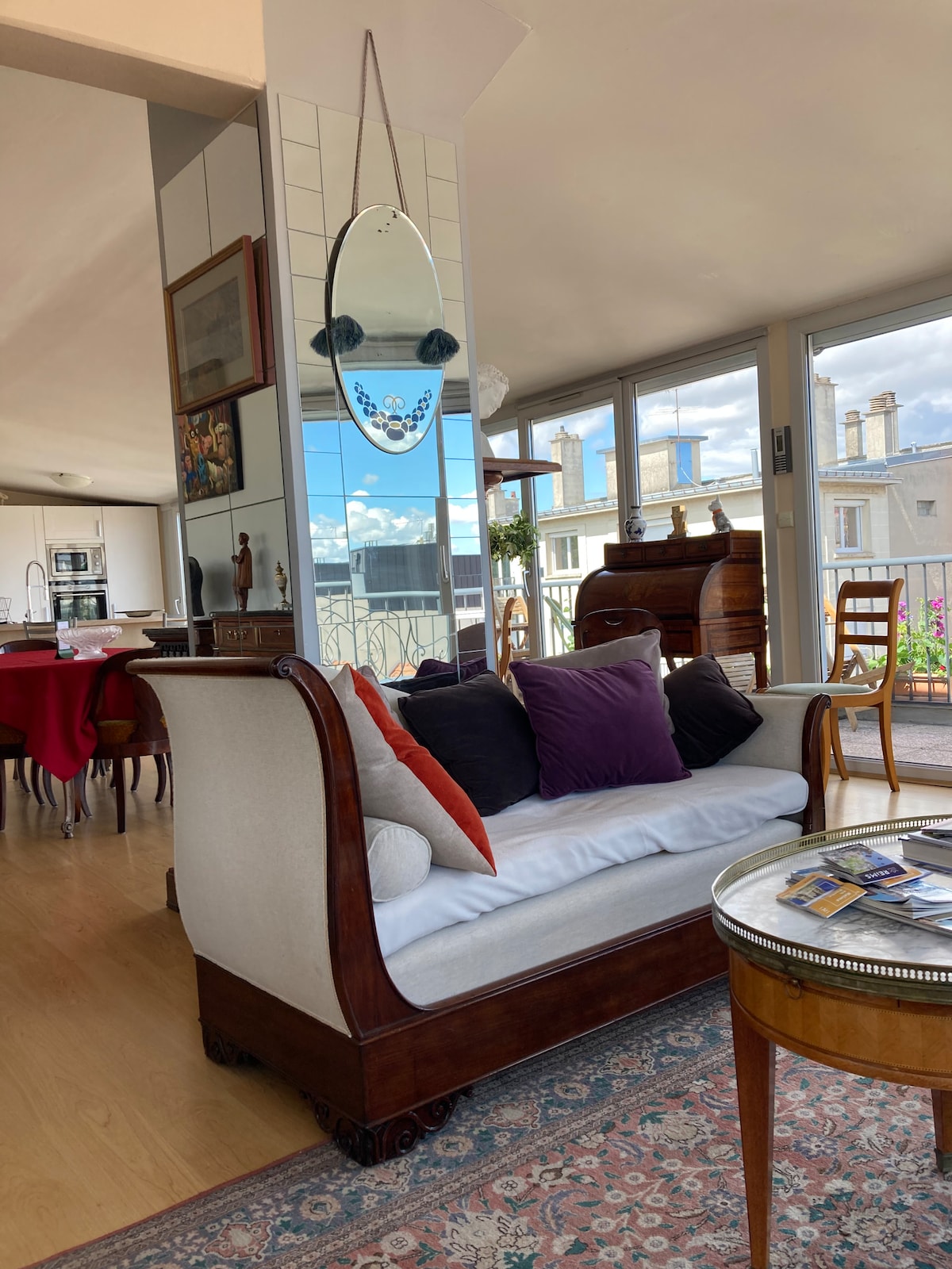 A comfortable seating area is featured with a stylish wooden sofa adorned with colorful pillows. A round side table sits nearby, while large windows provide ample natural light and showcase views of the surrounding rooftops. A decorative mirror hangs above the sofa.