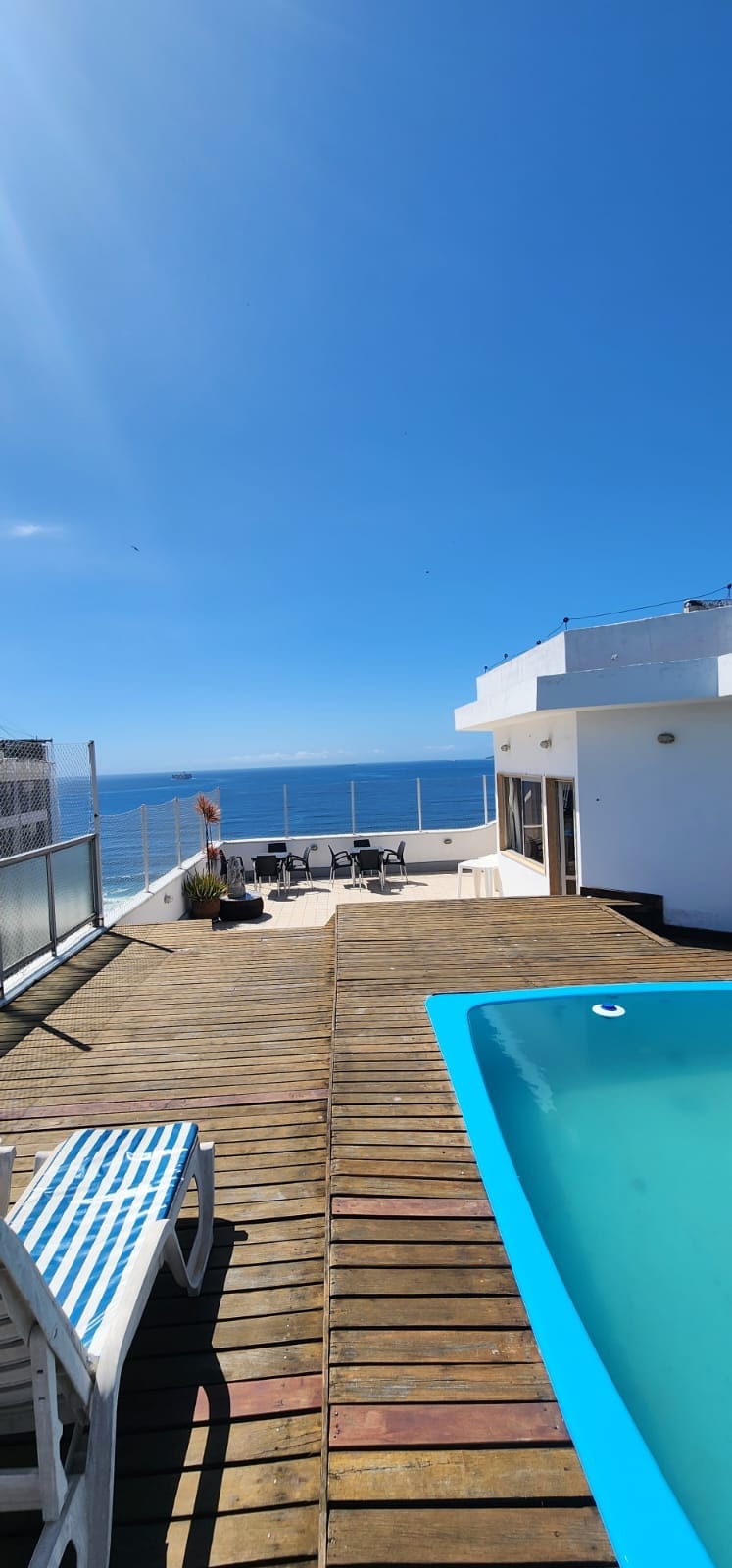 The image showcases a spacious deck with wooden flooring, adjacent to a private pool. Sun loungers are positioned nearby, and in the background, outdoor dining furniture is visible. The panoramic view features a clear blue sky and the shimmering sea, creating an inviting atmosphere.