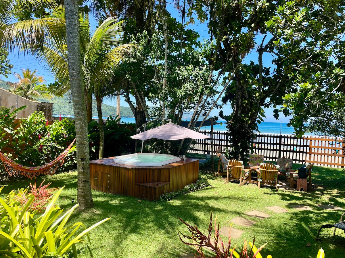 A serene garden area is surrounded by lush greenery and palm trees, featuring a wooden jacuzzi shaded by a pergola. Comfortable seating is arranged nearby, with a view of the ocean in the background, creating a perfect spot for relaxation.