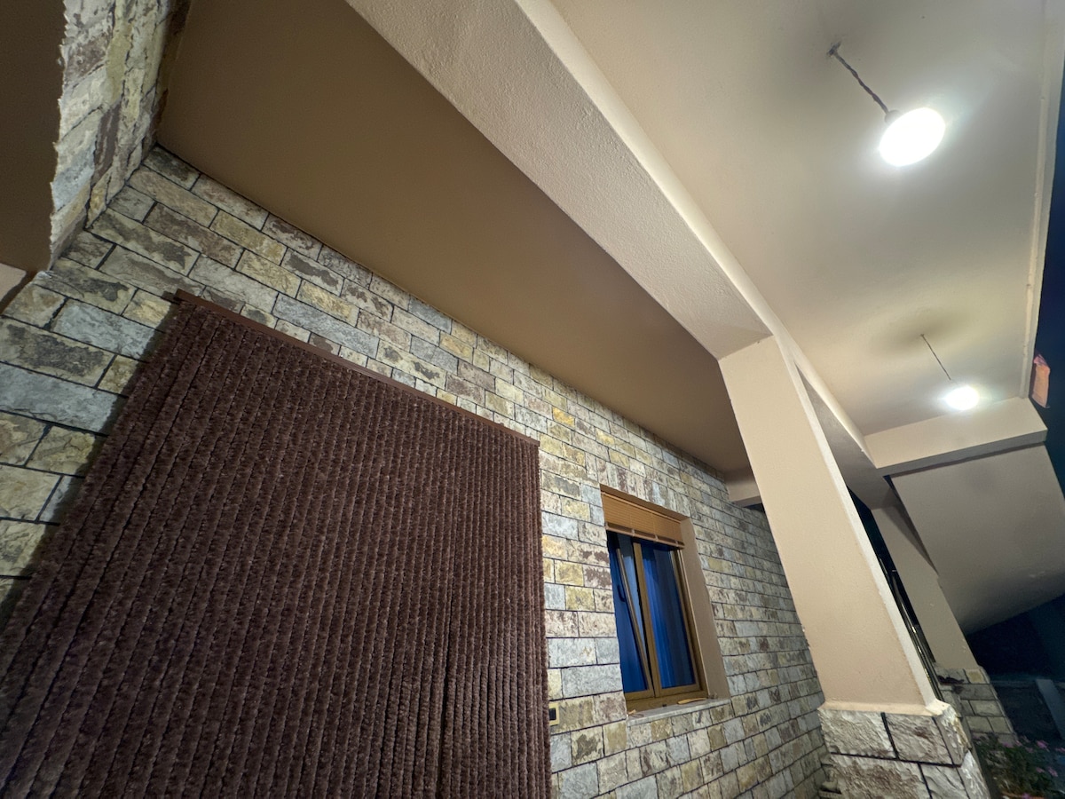 A well-lit exterior corner of a structure is displayed, featuring textured stone walls and a wooden window with blue curtains. A brown fabric wall hanging is visible, contributing to the welcoming appearance of the space.