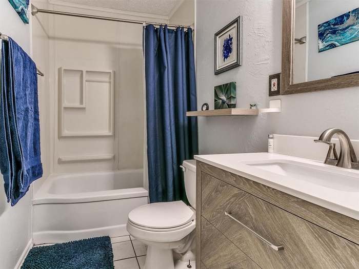 A streamlined bathroom features a bathtub with a shower curtain in a deep blue hue. A compact vanity with a storage cabinet stands beside a modern toilet. Textured towels hang on the wall, and a small shelf displays decorative items.