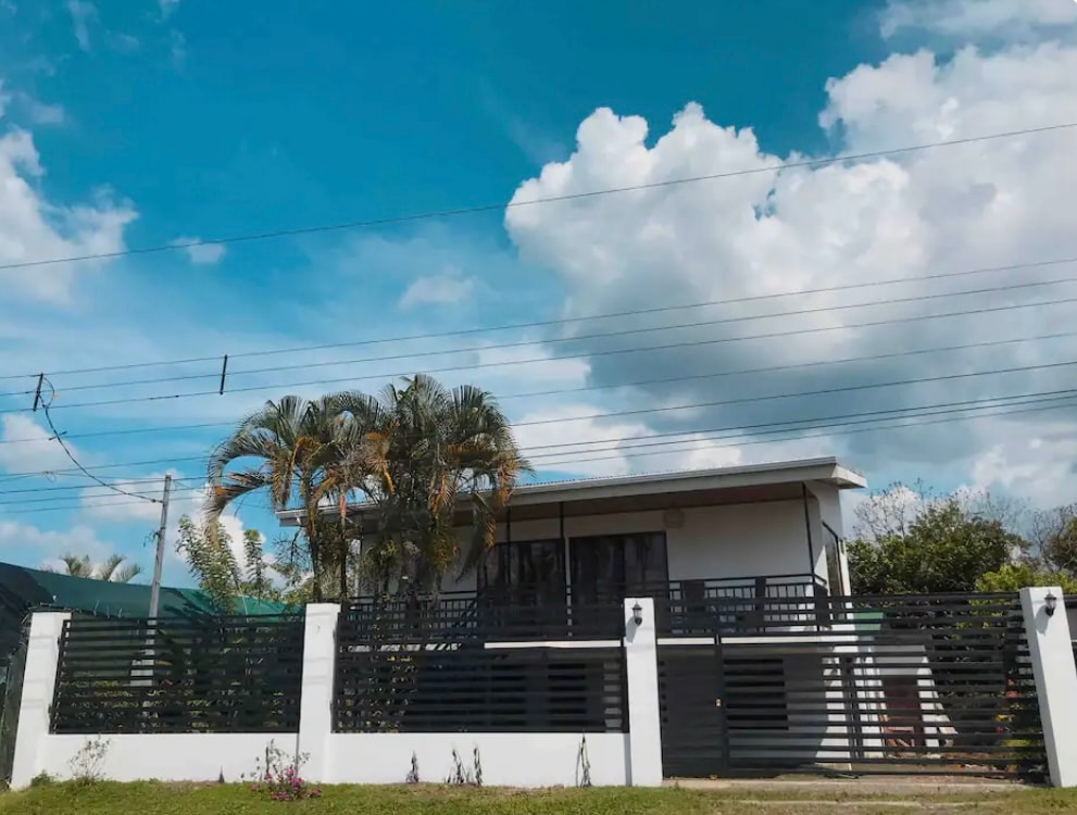 A modern building is surrounded by tropical greenery, featuring a secure front gate and a horizontal slatted fence. The sky above showcases fluffy clouds against a blue backdrop, creating a serene atmosphere.