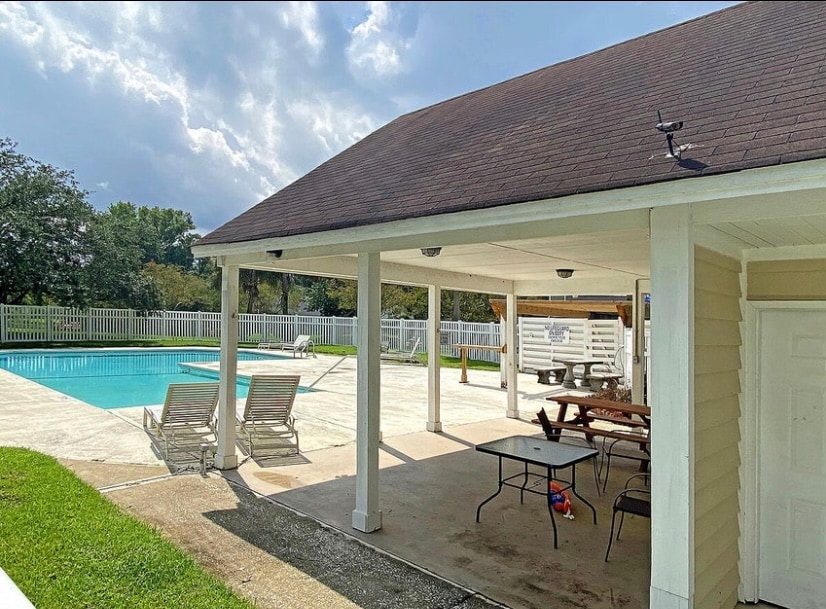 An outdoor area is shown, featuring a community pool surrounded by a concrete deck. Several lounge chairs are positioned near the pool, with a dining table and picnic bench located under a covered seating area. Manicured grass borders the space.