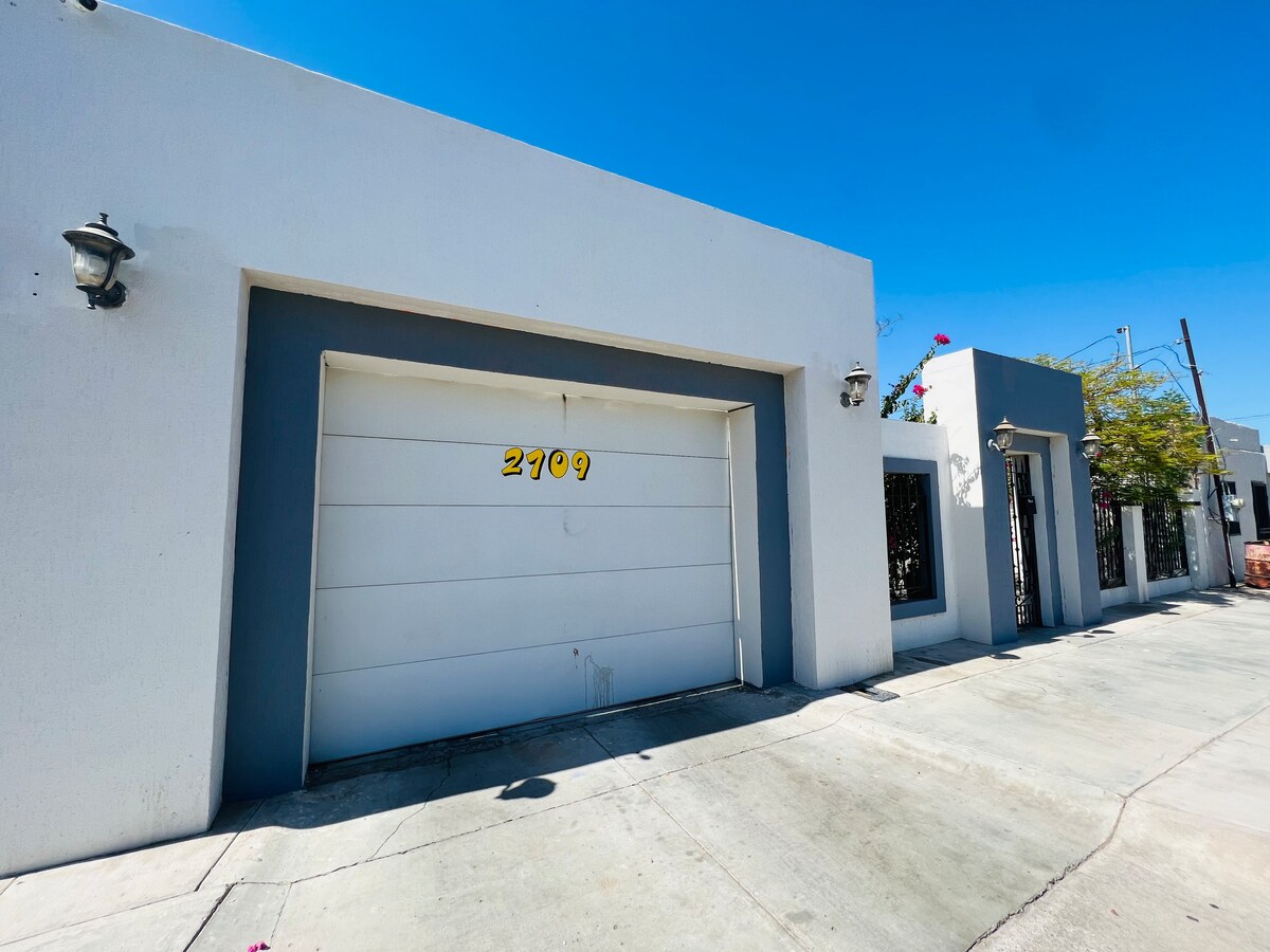 A modern white house exterior features a large garage door with the number 2709 clearly displayed. The walls are painted in contrasting grey and white, complemented by decorative accents. Bright sunlight highlights the clean lines of the structure.