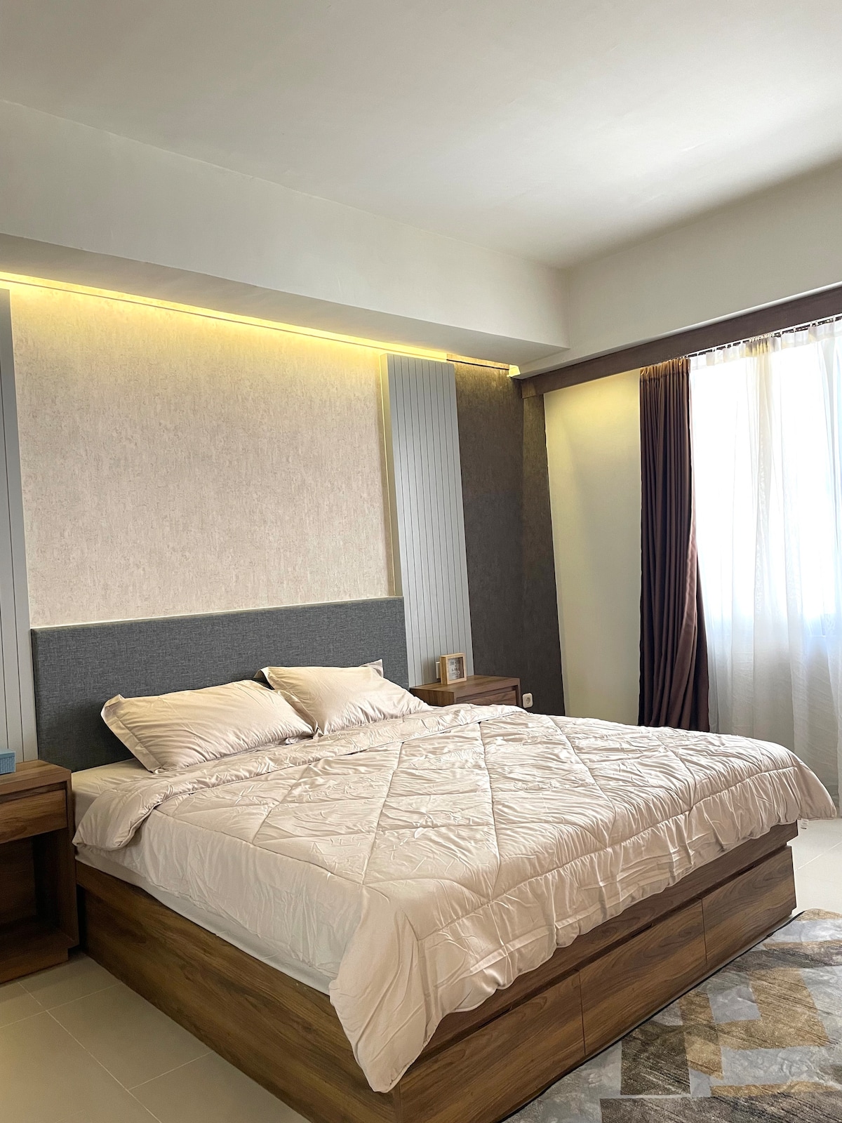 A spacious sleeping area features a queen bed adorned with a light-colored quilt. Soft lighting highlights the textured wall behind the bed, while a large window, dressed in flowing curtains, allows natural light to fill the room.