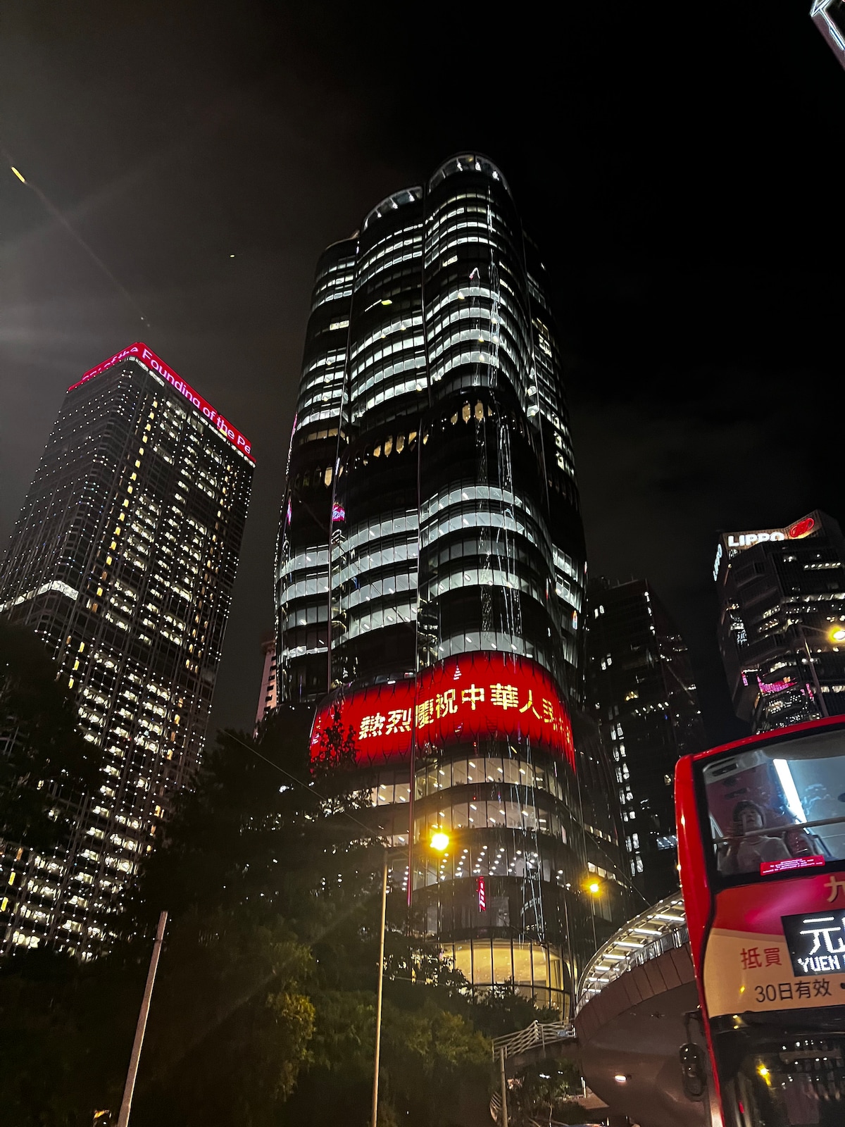 A tall modern building reaches towards the night sky, featuring a sleek glass exterior that reflects surrounding city lights. Bright red advertisements illuminate the façade, creating a vibrant urban atmosphere. Nearby street-level elements include vehicles and greenery, emphasizing the bustling city environment.
