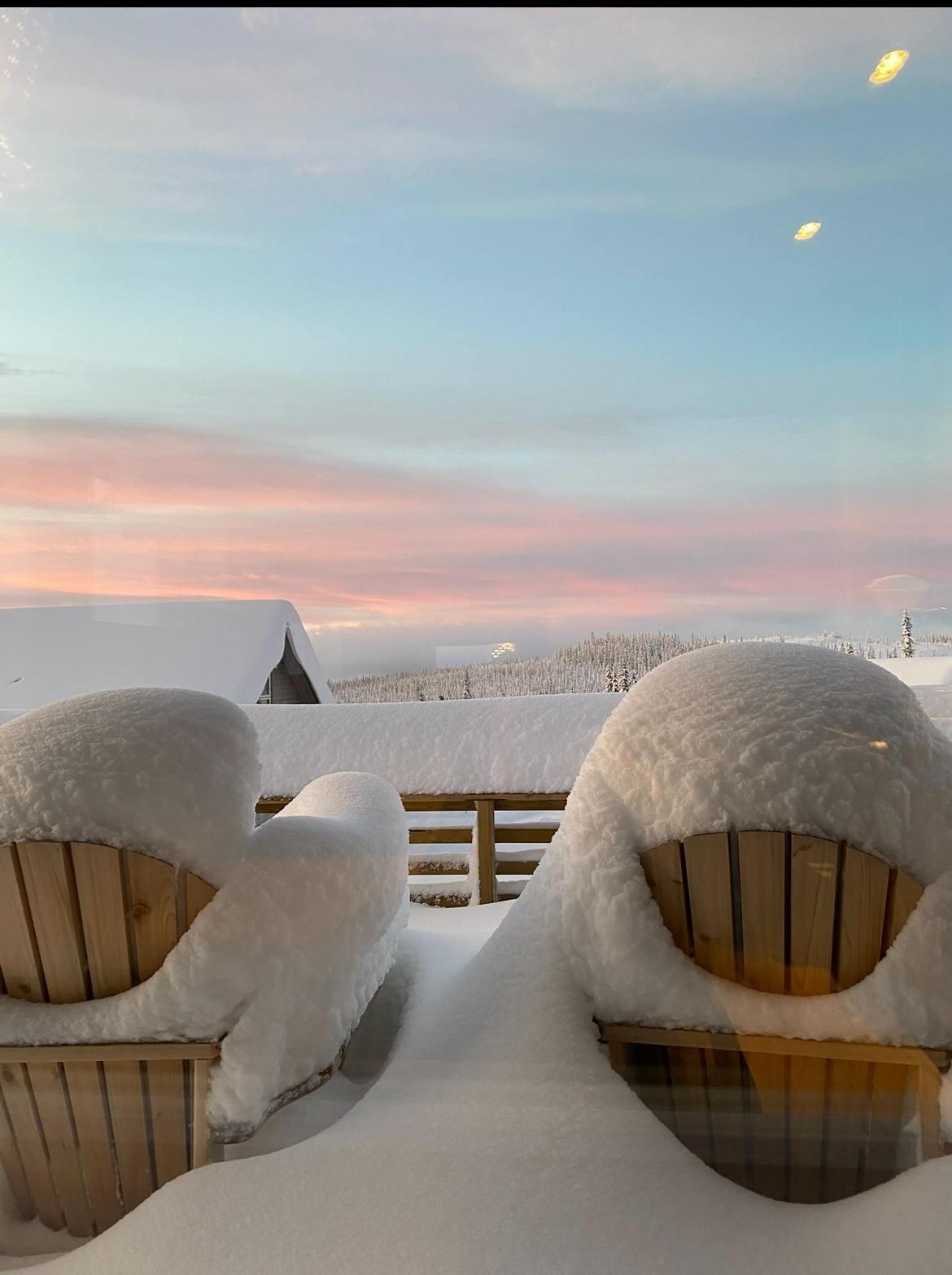 Two wooden chairs are covered in fresh, soft snow on a snowy terrace. A picturesque view reveals a serene landscape, with gentle slopes and trees blanketed in white. The sky transitions from light pink to soft blue, indicating a calm, wintry evening.