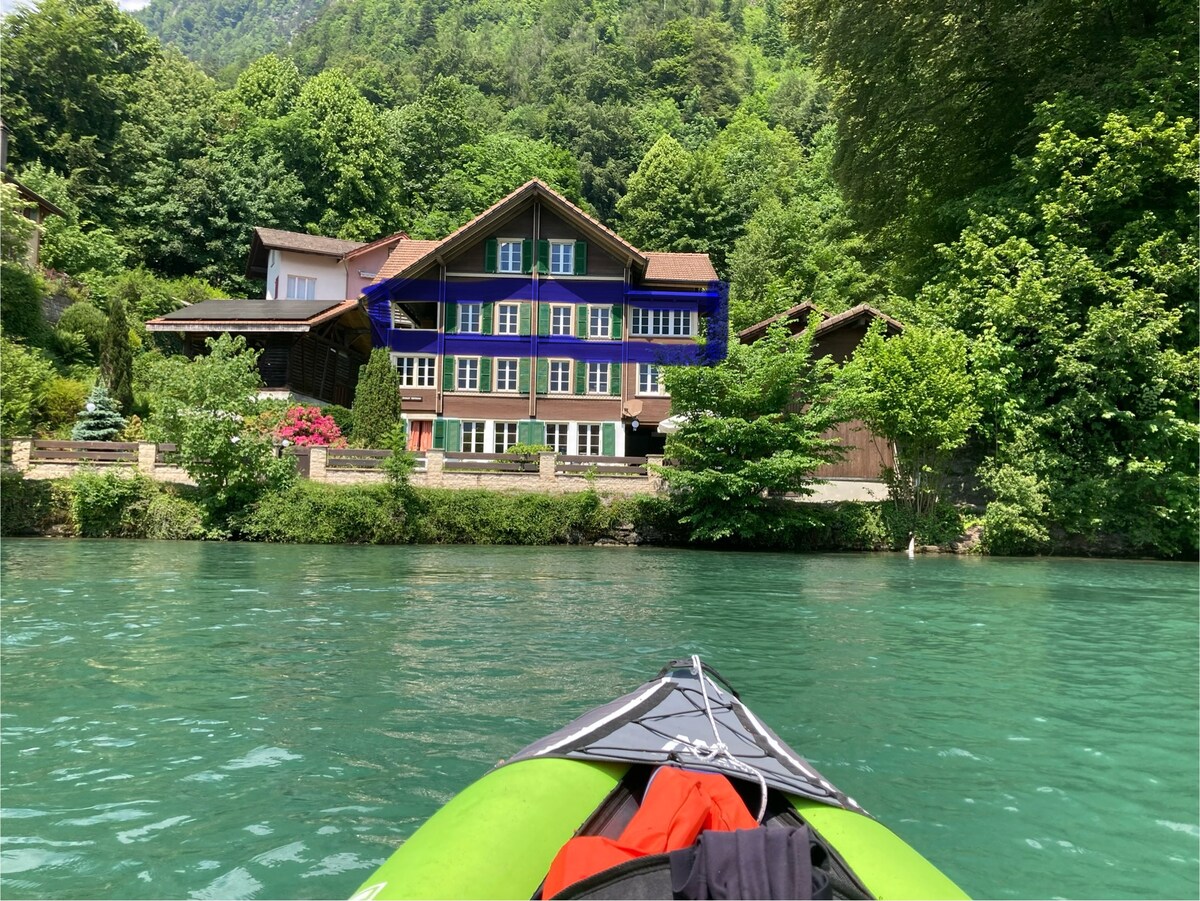 A large, multi-story building is situated along the banks of a turquoise river, surrounded by lush green trees and plants. The structure features multiple windows, providing a view of the natural landscape. A kayak is partially visible in the foreground, hinting at nearby water activities.