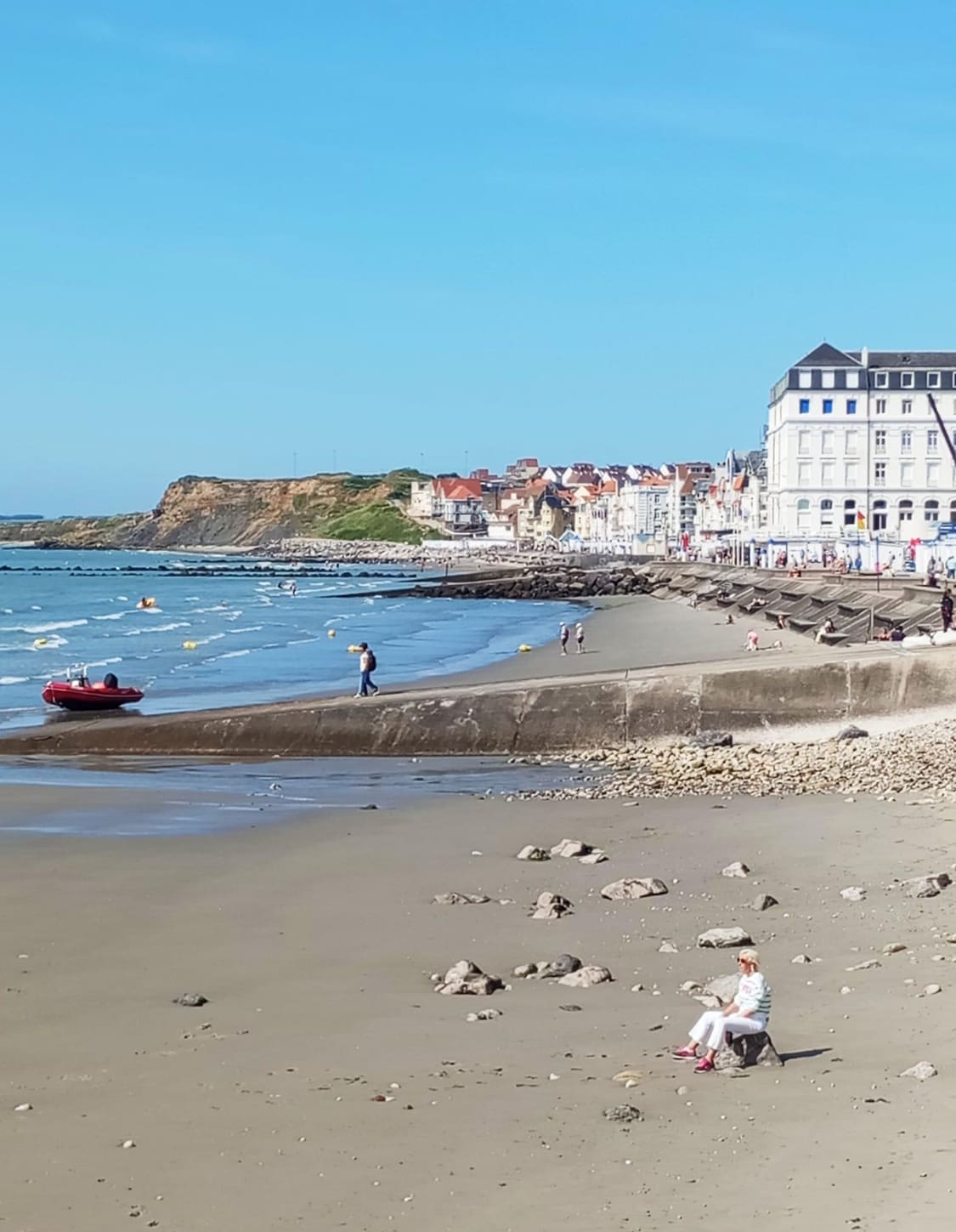 The beach scene captures a shoreline lined with smooth sand and rocky formations. Calm waves lap against the coast, where individuals stroll along the water's edge. A small boat rests near the beach, while colorful buildings are visible in the background against a clear blue sky.