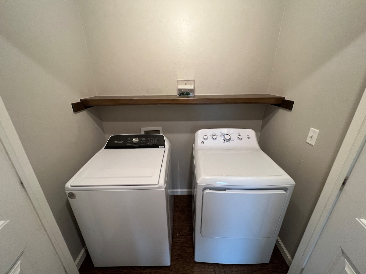 A dedicated laundry room is equipped with a black washing machine and a white dryer, positioned side by side against a neutral wall. Above, a wooden shelf provides additional storage space, allowing for a tidy and functional laundry area.