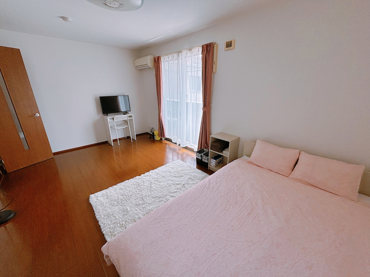 A bright and airy bedroom features a spacious bed dressed in light pink bedding. Natural light enters through the sheer curtains, illuminating the wooden floor. A small desk and a television are positioned against the wall, while a cozy white rug adds comfort to the space.