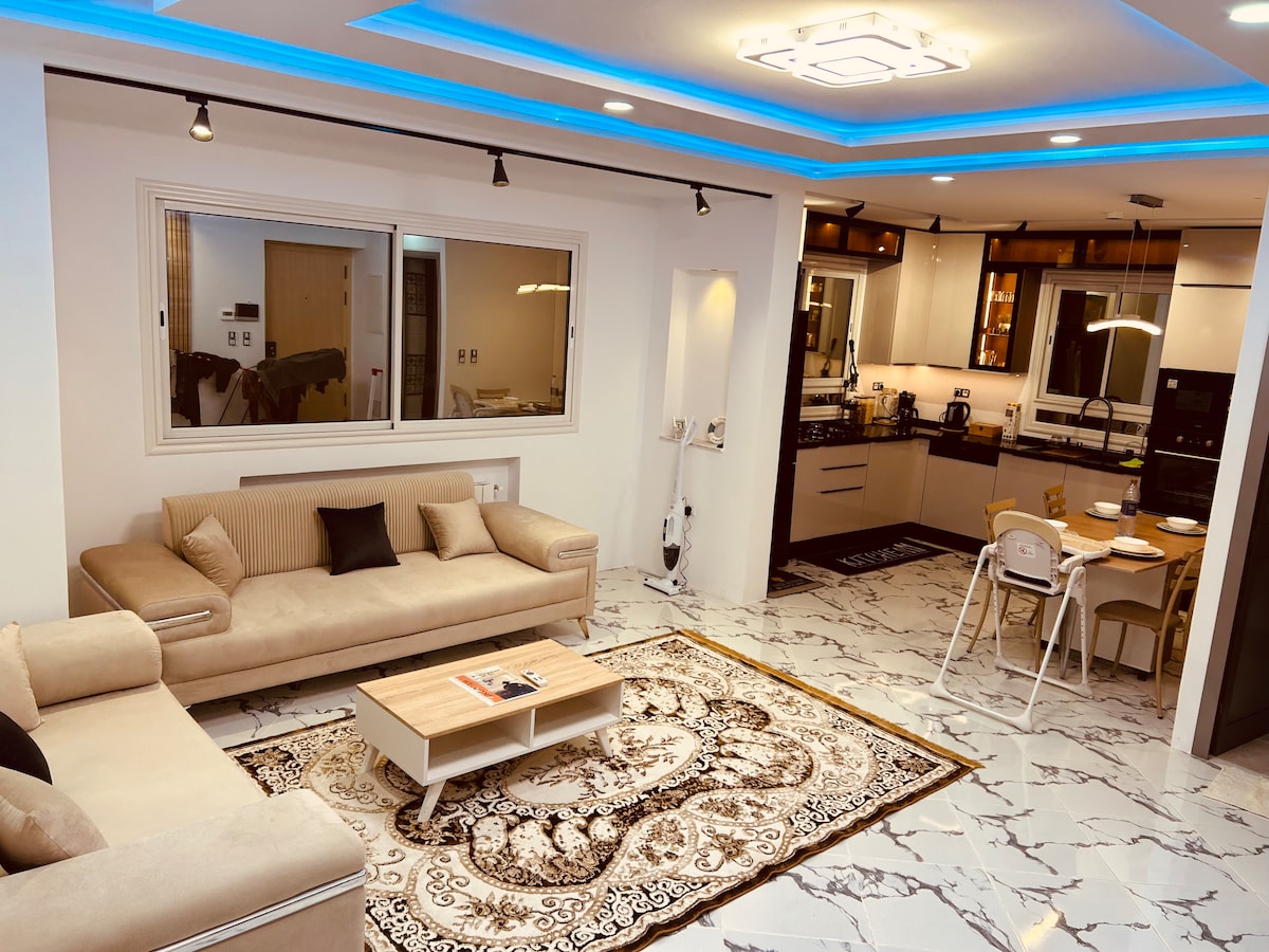 A spacious living area features a light-colored sofa set facing a decorative coffee table. A patterned rug anchors the space, while large windows provide natural light. The modern kitchen is visible, equipped with sleek cabinetry and dining space for two.