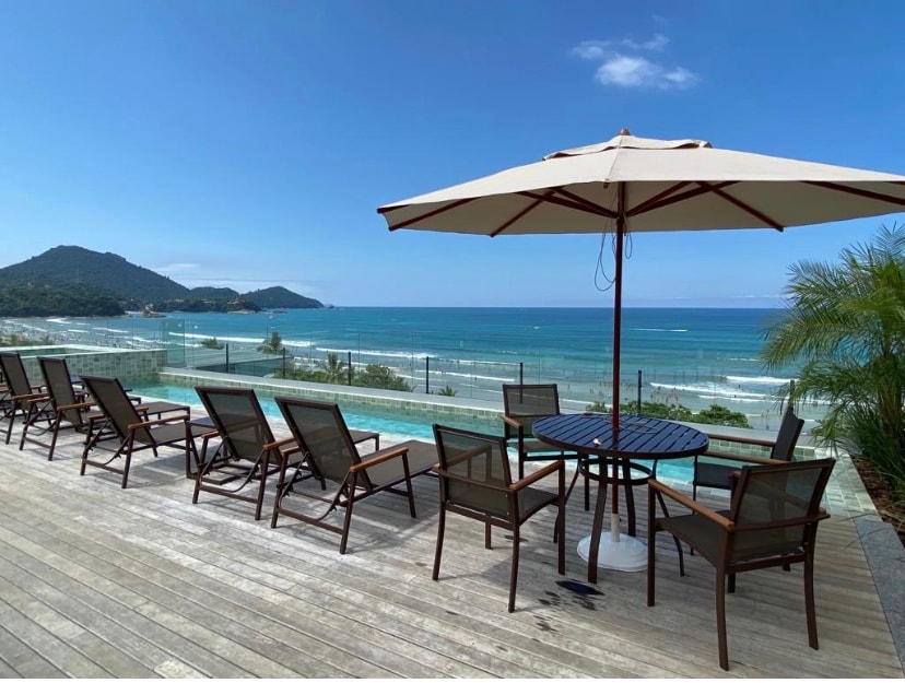 A spacious outdoor deck features multiple brown lounge chairs arranged around a circular table. An umbrella provides shade, overlooking a scenic view of the ocean and distant hills. The tranquil infinity pool blends with the coastline, enhancing the picturesque environment.