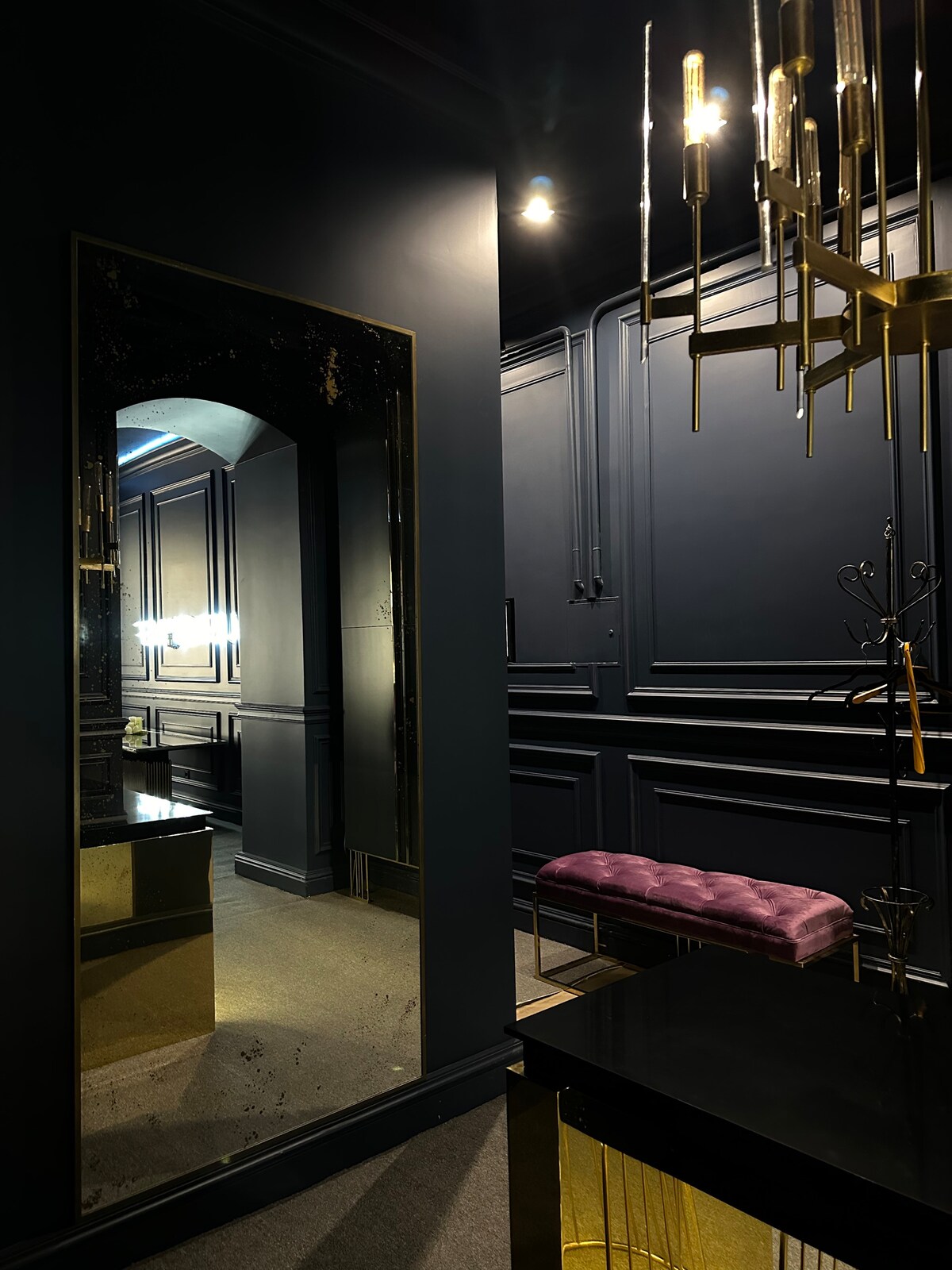 A stylish entryway is featured, showcasing dark walls with elegant gold accents. A large mirror reflects the space, while a plush purple bench is positioned on a black surface. Soft lighting illuminates the area, enhancing the sophisticated design.