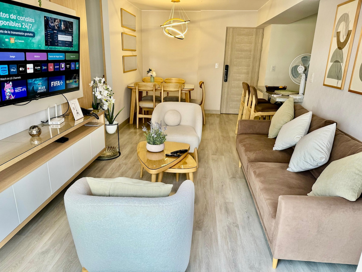 The living area features a neutral palette with a brown sofa and light-colored armchairs. A modern entertainment unit holds a large TV displaying various channels. A dining table with chairs is visible in the background, and a ceiling light adds a contemporary touch to the space.