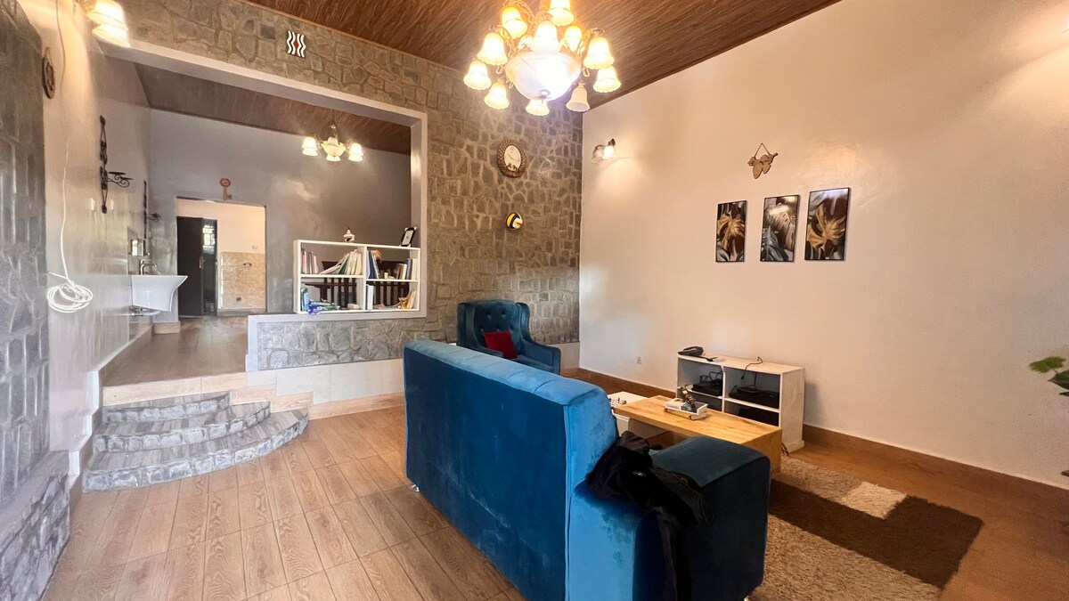 A stylish living area is highlighted by a blue sofa and wooden coffee table. Stone walls provide a natural texture, while a few decorative light fixtures enhance the ambiance. An open space is visible in the background, integrating additional living arrangements.
