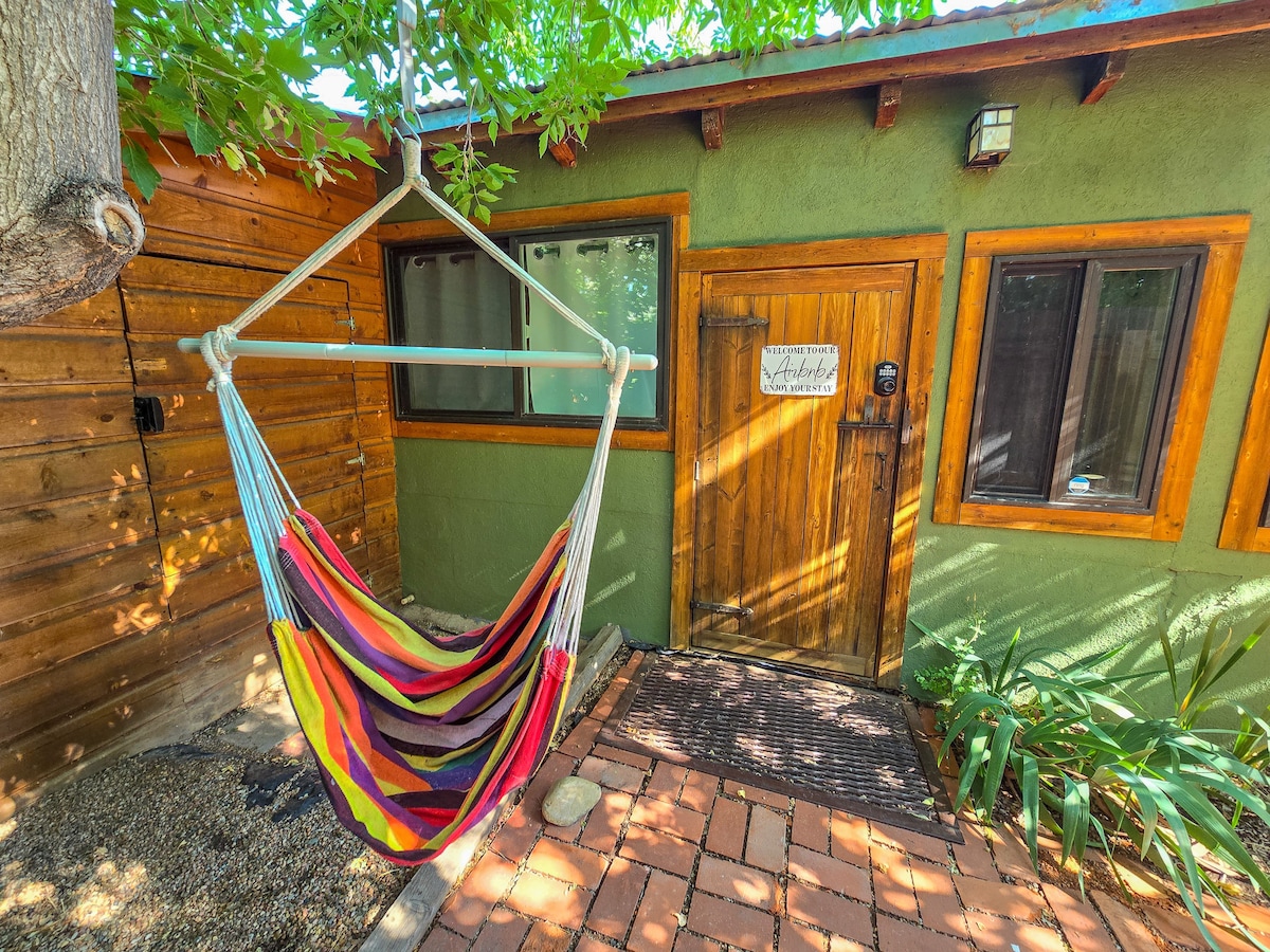 A vibrant hammock is suspended near the entrance of the Casita, inviting relaxation. The wooden door features a decorative sign, while surrounding windows allow natural light to enter. Lush greenery frames the entrance, contributing to a tranquil atmosphere.