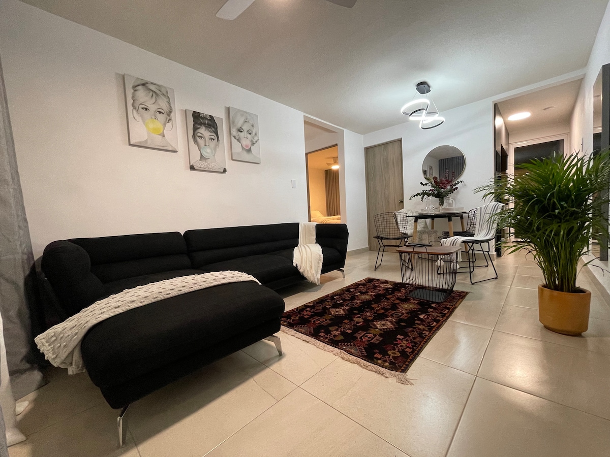 A modern living area is presented with a sleek black sectional sofa and a decorative area rug. A dining table with four chairs is situated nearby, accentuated by a plant and wall art above the sofa. Soft lighting adds warmth to the inviting space.