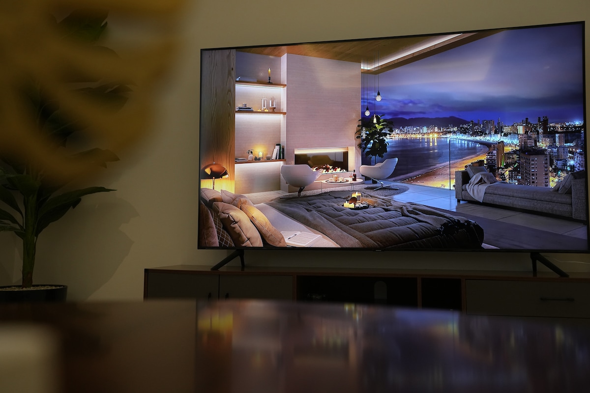A modern living space is featured on a large television screen, showcasing a serene city view at dusk. The image highlights a cozy seating area with plush blankets and warm lighting, framed by contemporary decor and a panoramic window overlooking the water.