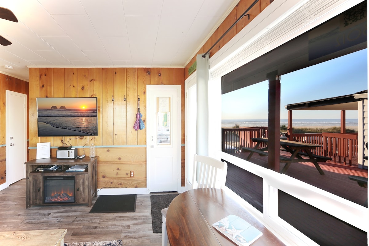 A cozy living area features warm wooden paneling and a large window offering a clear view of the horizon. A TV is mounted on the wall, complemented by a stylish media console. A round dining table is positioned nearby, inviting shared meals and conversation.