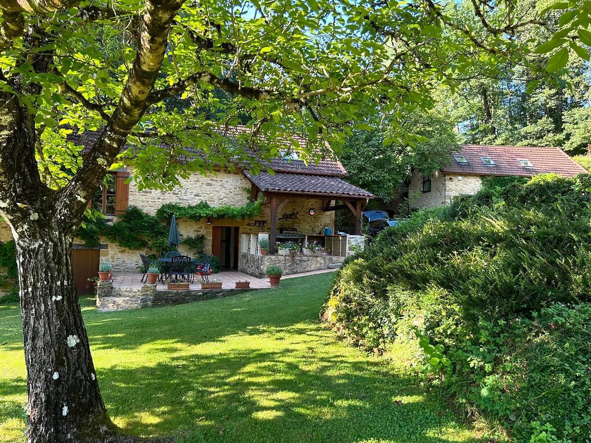 Two stone cottages are nestled among lush greenery, with the larger cottage featuring a shaded terrace adorned with potted plants. The inviting outdoor space includes a well-kept lawn and a backdrop of trees, creating a serene atmosphere for relaxation.